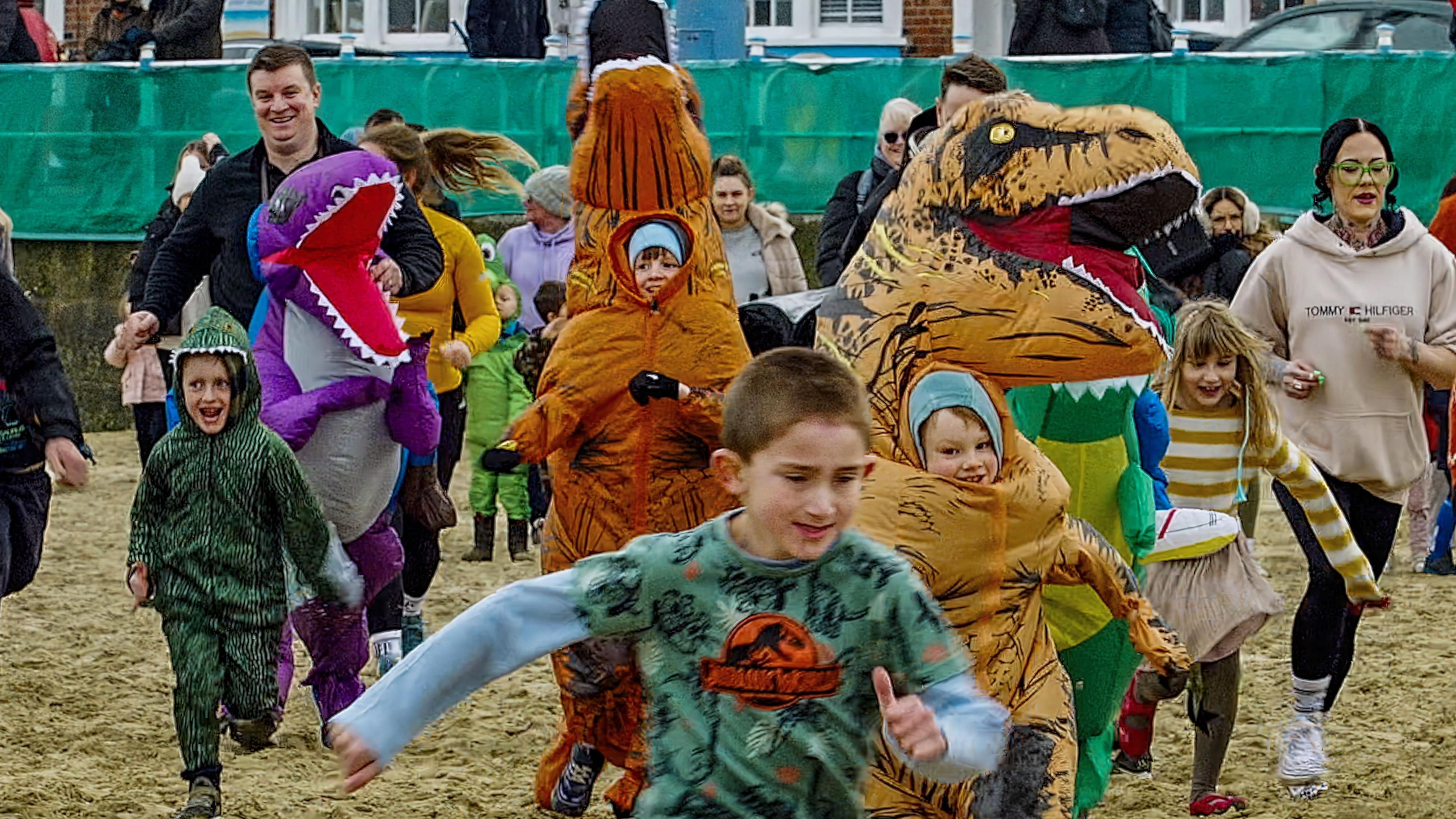 A group of children and adults wearing dinosaur costumes running along a beach.