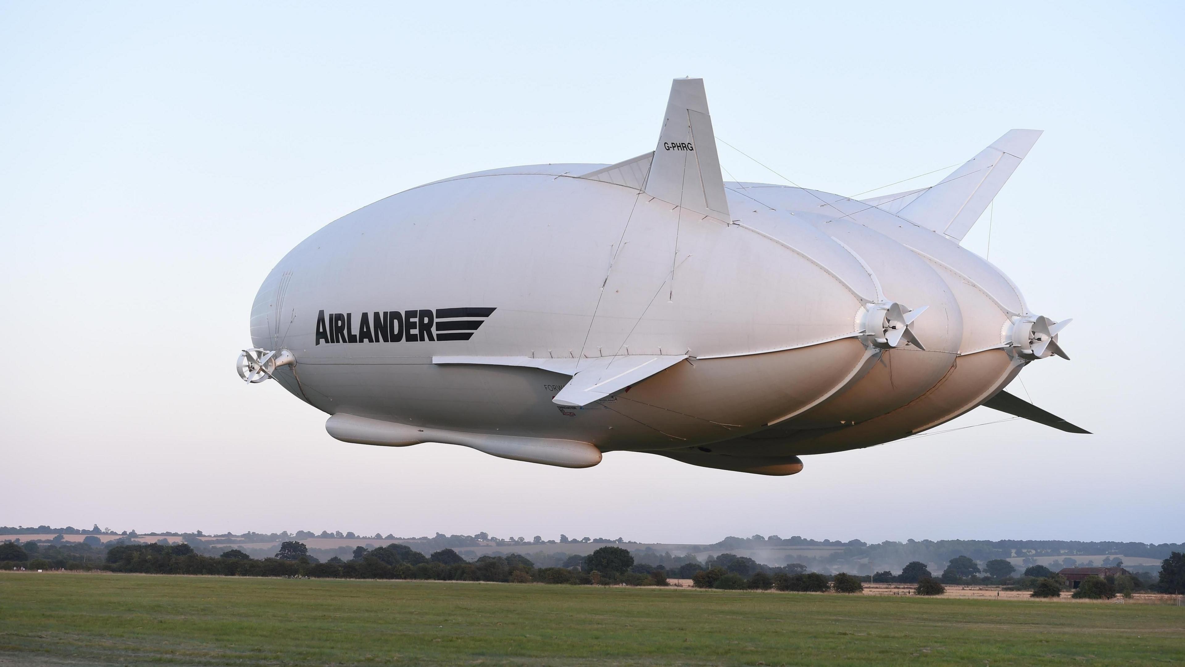 A large airship which is floating in the air above green fields. It has two small propellers at the back and on the side. The word "Airlander" is written in black on the side.