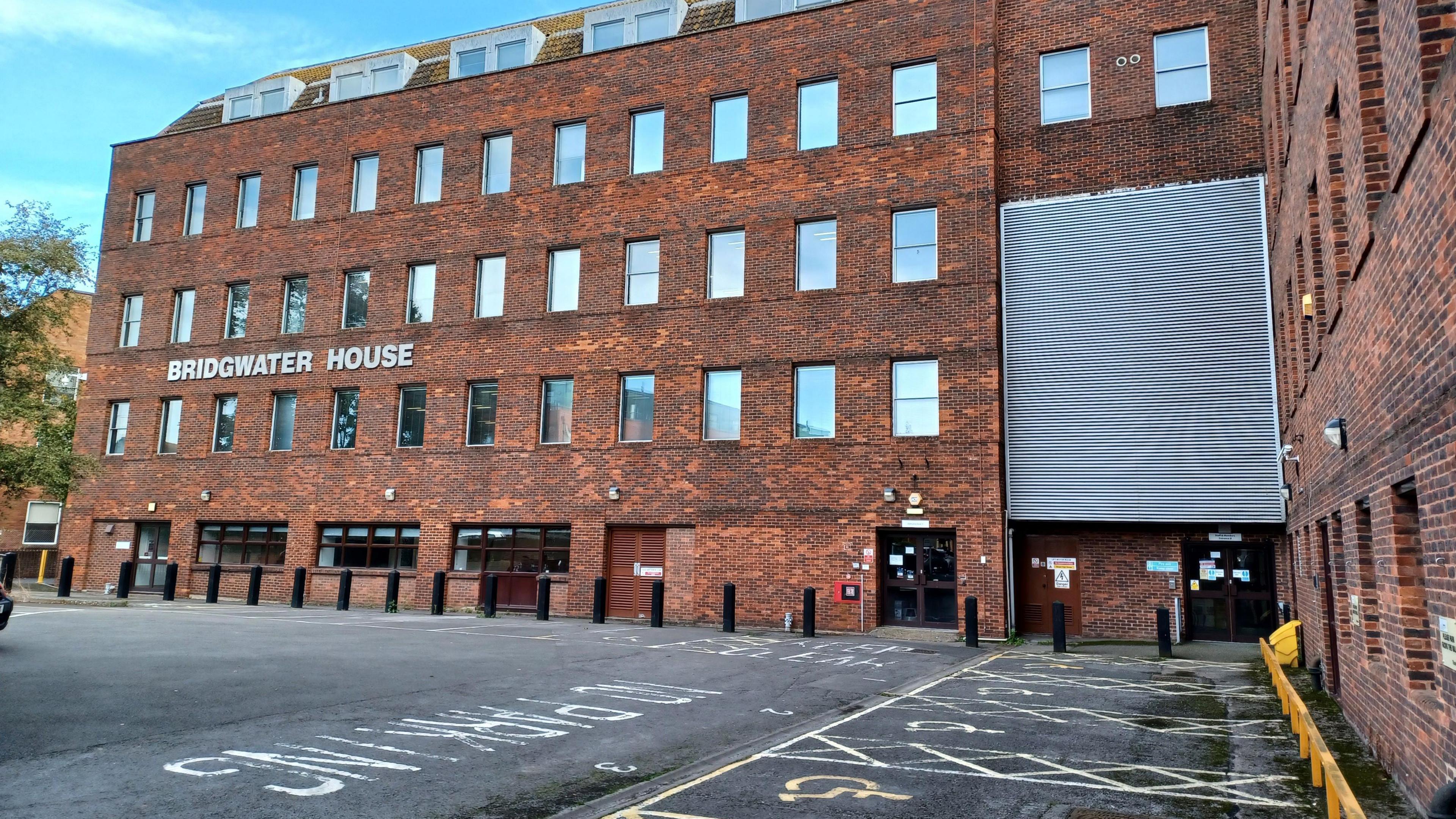 Car park with red brick building with a large grey shutter covering multiple floors. The building has "Bridgwater House" in large pale letters between the first and second floor