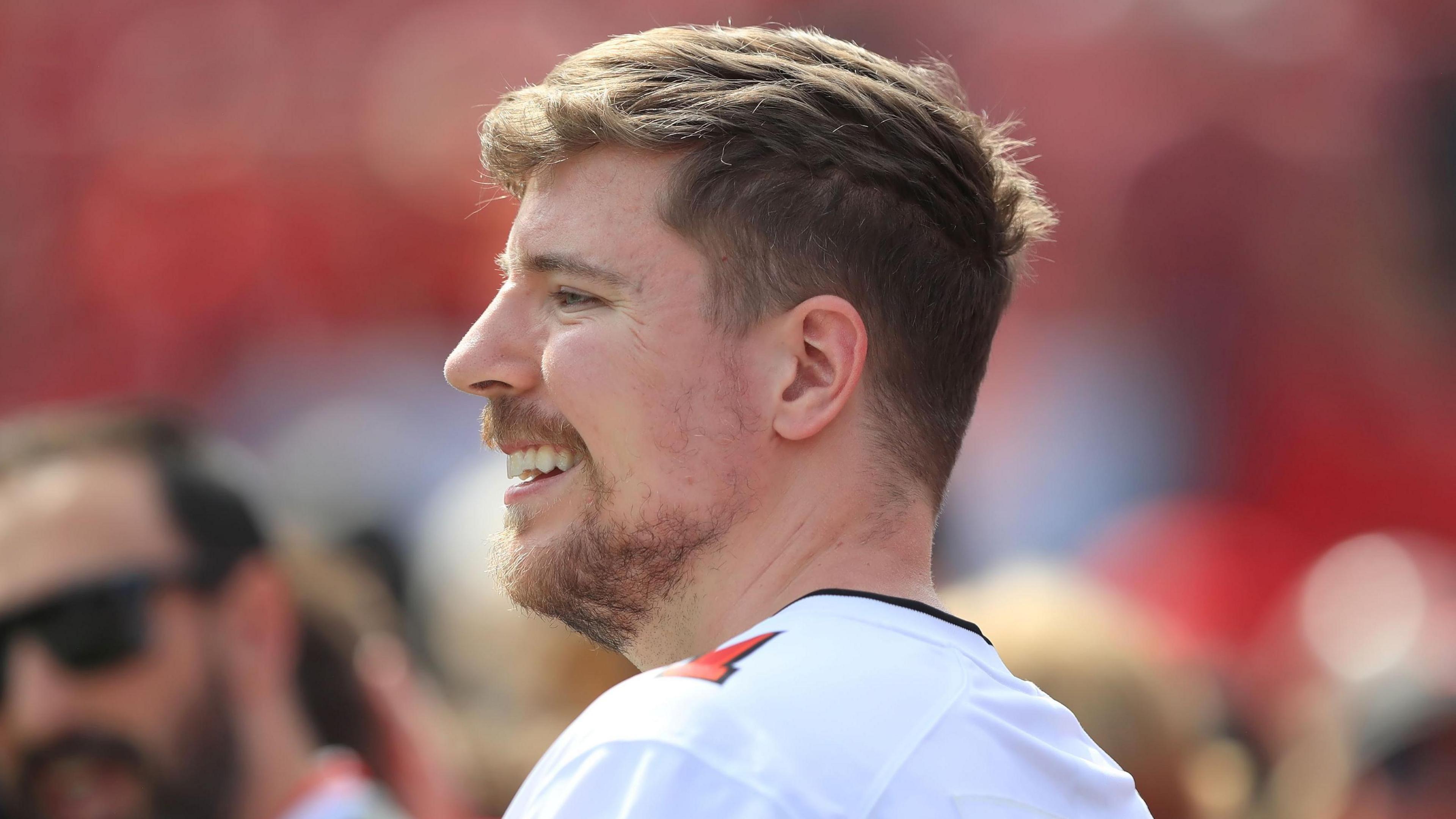 MrBeast AKA Jimmy Donaldson pictured at an American Football game. He wears a white football shirt with red numbering on his shoulder. He has a short beard and short sandy hair. 