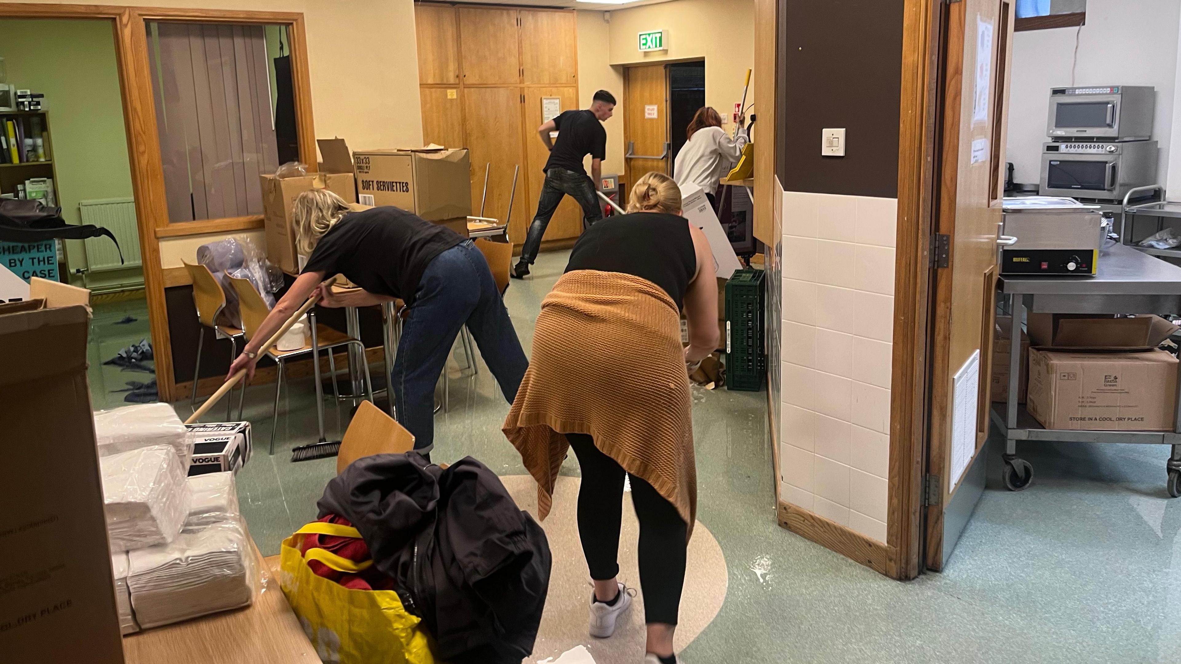 Volunteers seen mopping up flood water in the cafe space. There are four volunteers visible and the room is messy, with boxes and supplies lifted on to tables and chairs stacked up. Blue paper soaked with water can be seen on the floor. 