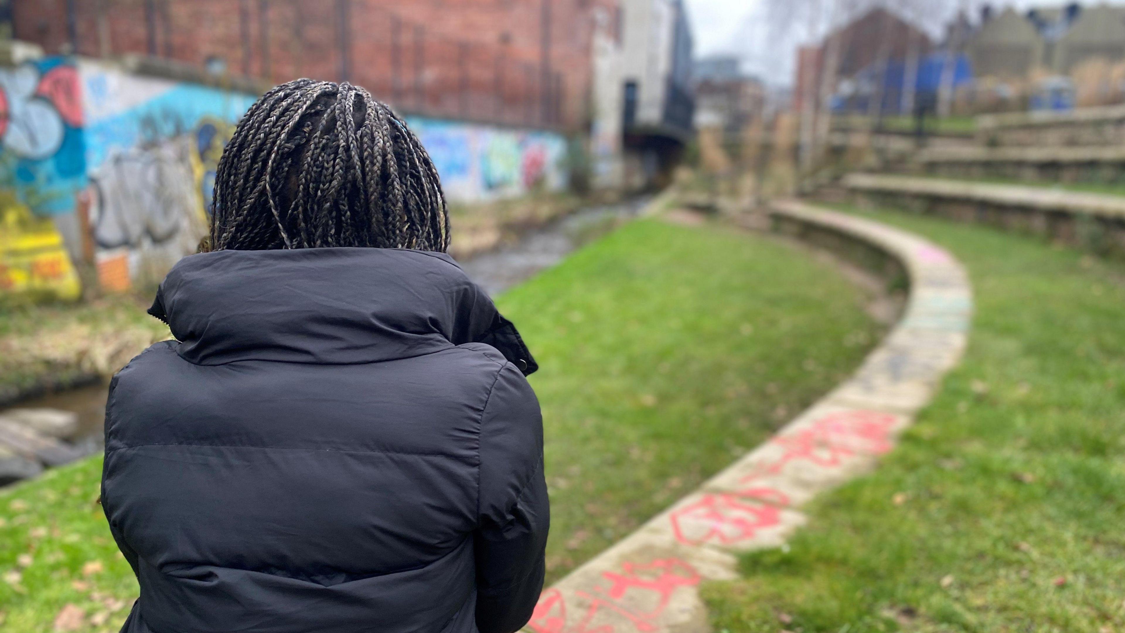 A woman wearing a black coat stands with her back to the camera, in front of a stream with graffiti on a wall in the background.