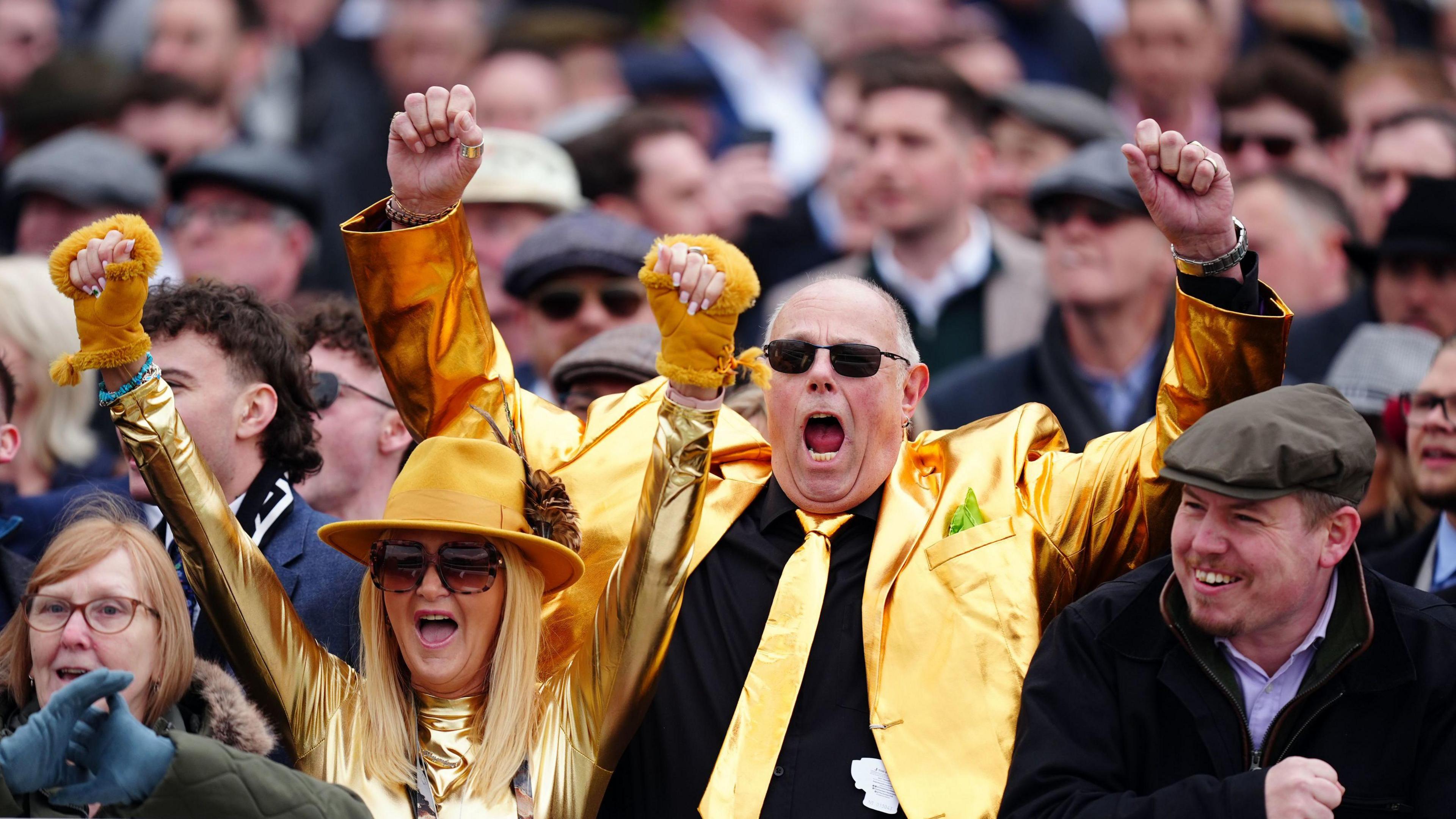 A man and a woman wearing shiny gold outfits cheer for joy with their arms raised in the air. They are in a crowd of people who are eagerly watching the races.