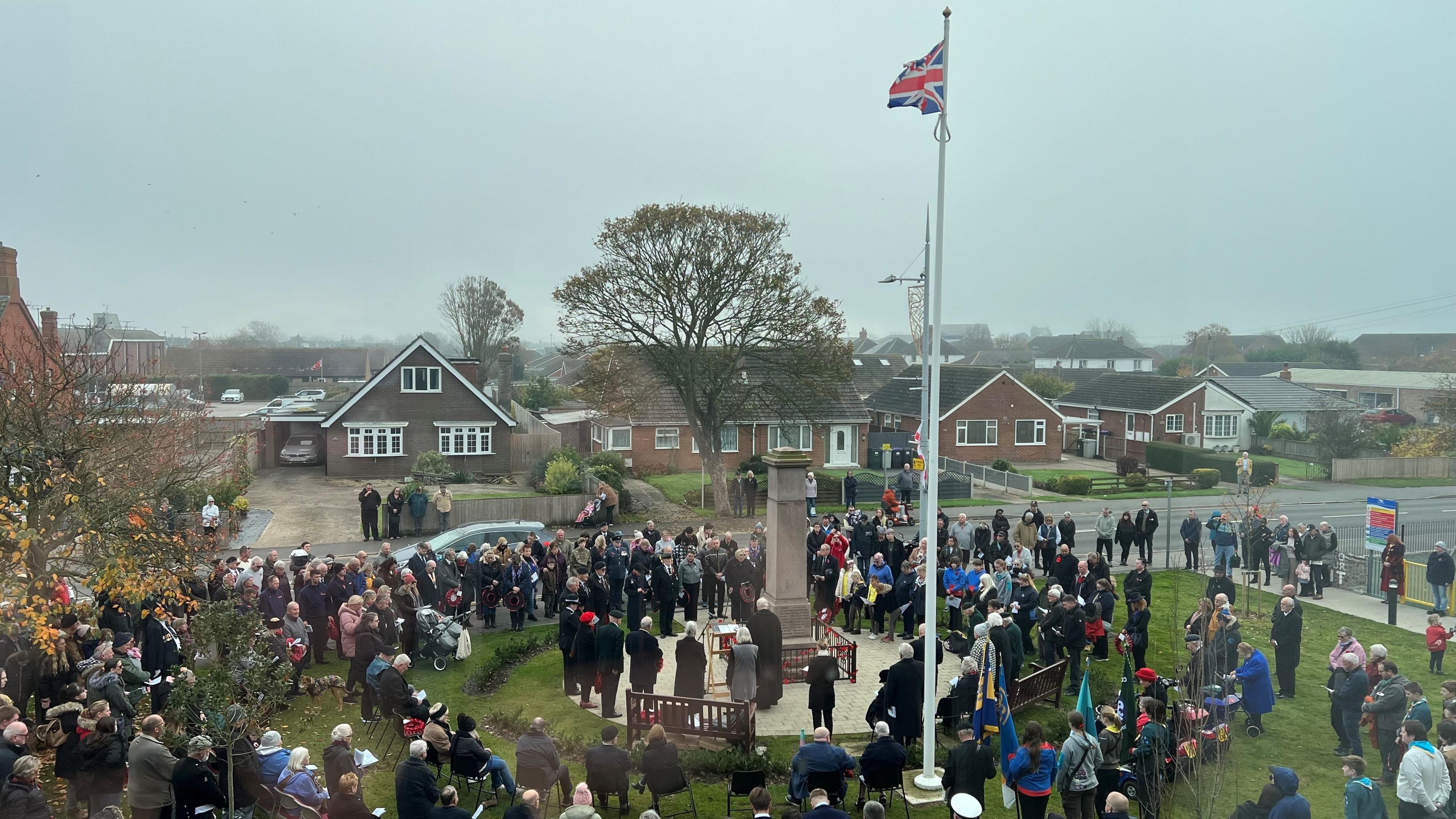 A photo of crowds gathered at the memorial on a grey day. The union jack flag flies on a tall pole above the memorial.