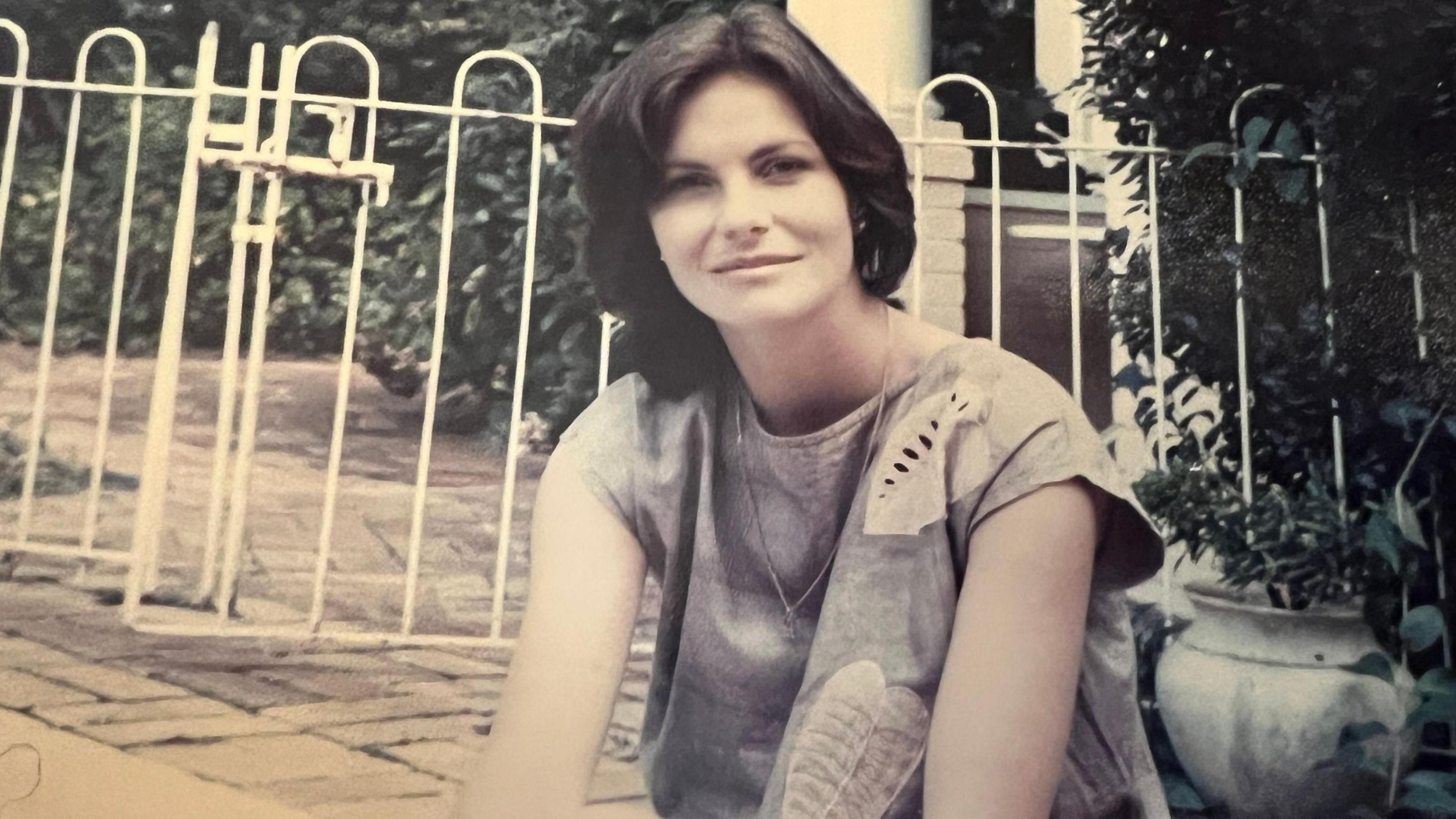 The picture shows a person sitting outdoors near a white metal fence. They are wearing a short-sleeved shirt with a leaf pattern and have short hair. In the background, there are plants and greenery, and a large ceramic pot is visible on the right side. 