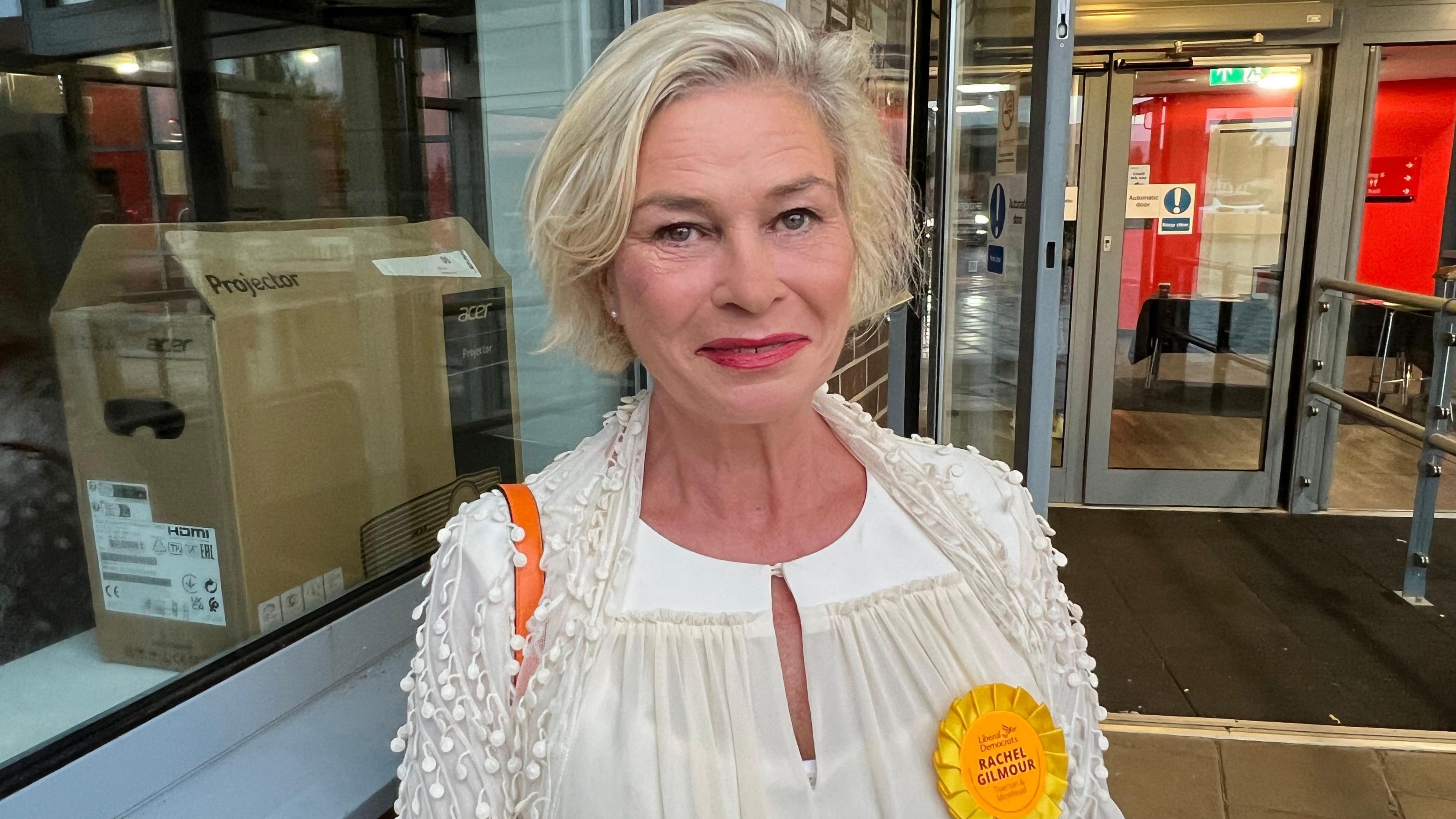 Image of Rachel Gilmour. She has blonde short hair and is wearing a white blouse and a yellow Liberal Democrat badge 