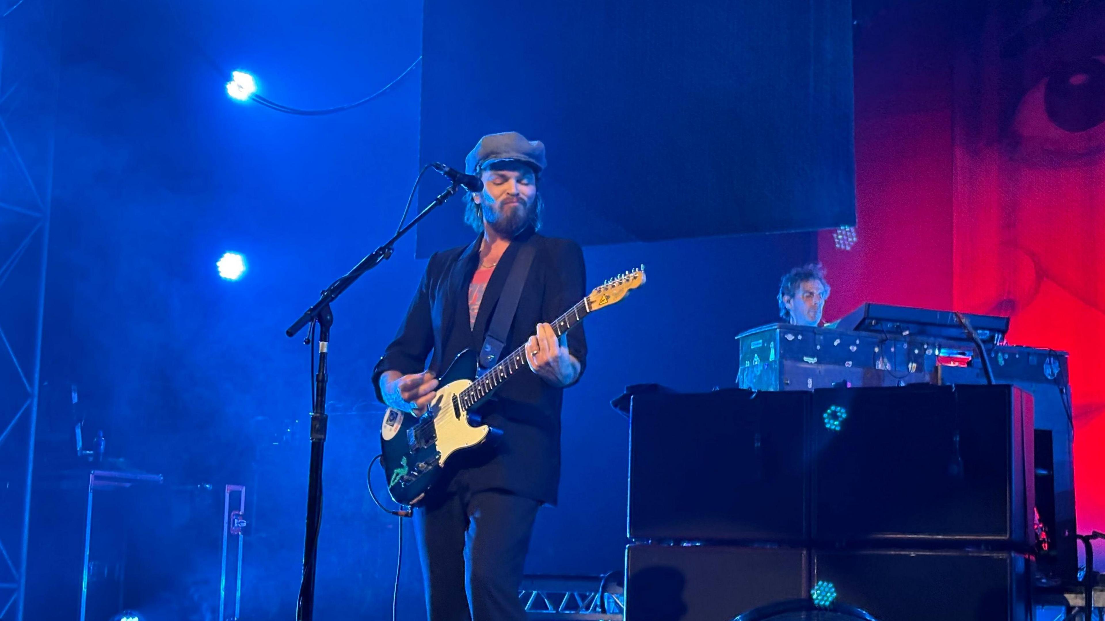 Gaz Coombes has a dark beard and medium length dark hair. He wears a flat cap and a dark suit. He is playing a guitar.