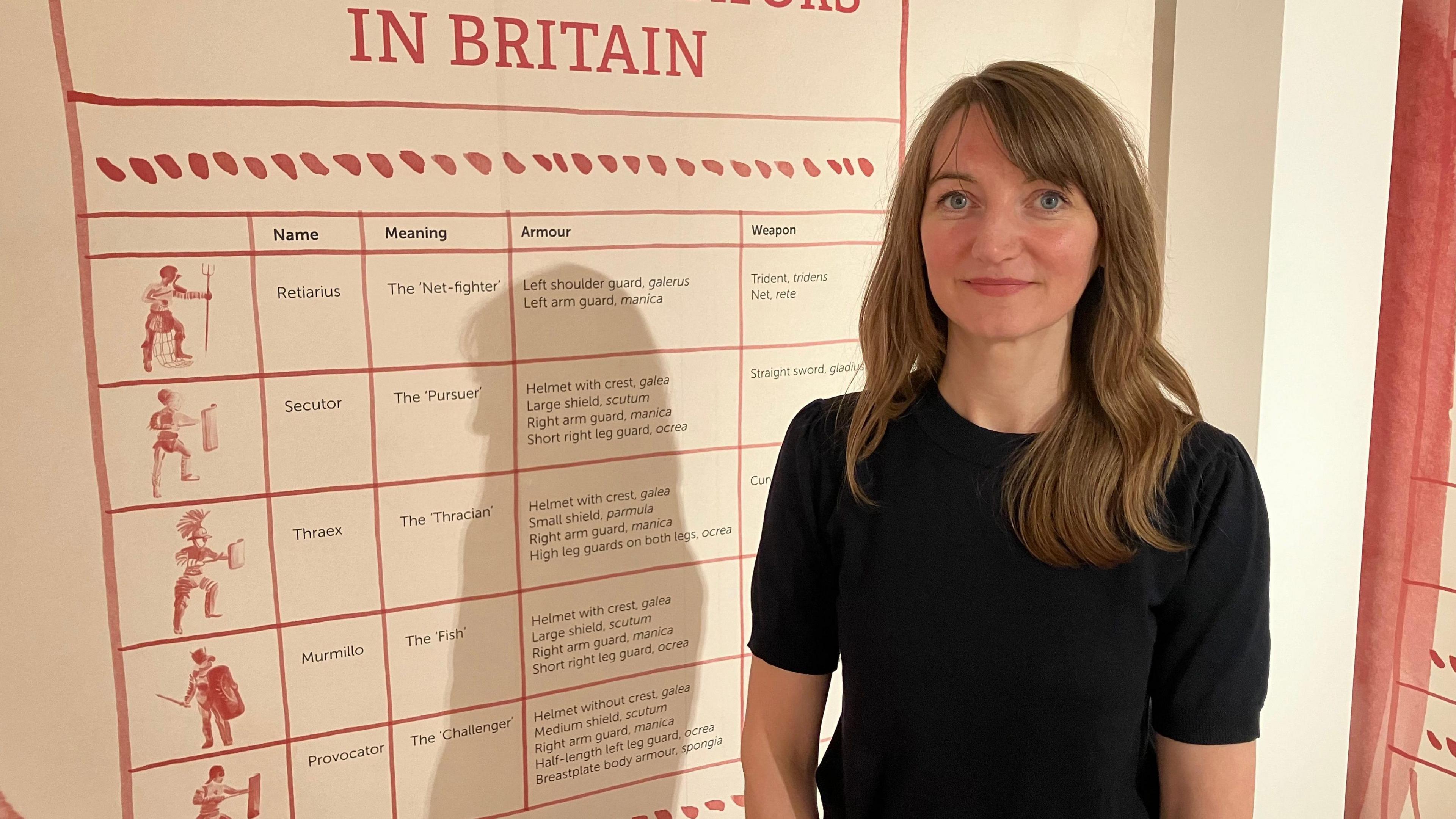 A woman in a black dress stands next to a wall chart showing different types of gladiators.