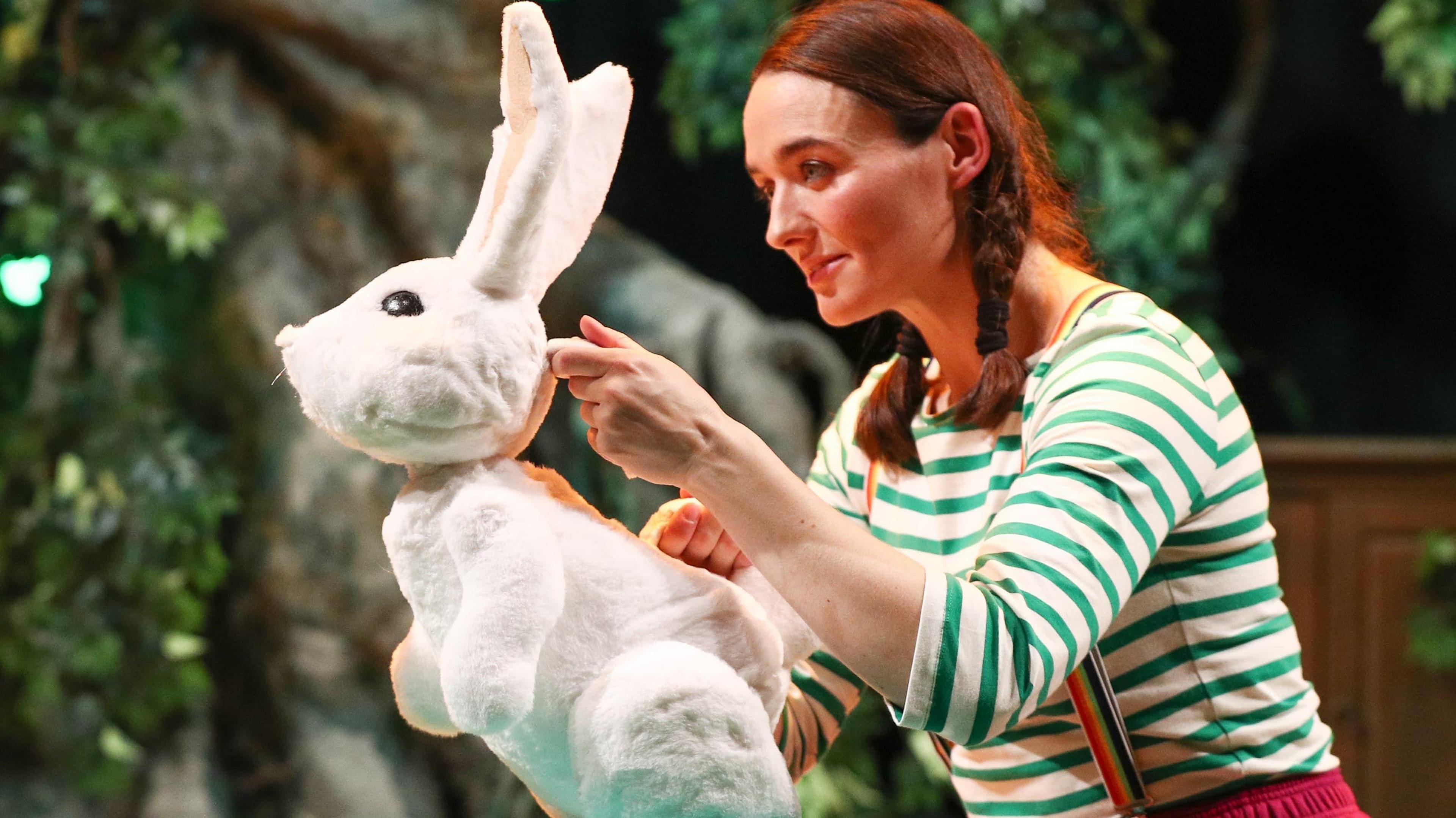 Anna holding a white rabbit puppet during a performance wearing a striped top and auburn hair in pigtails.