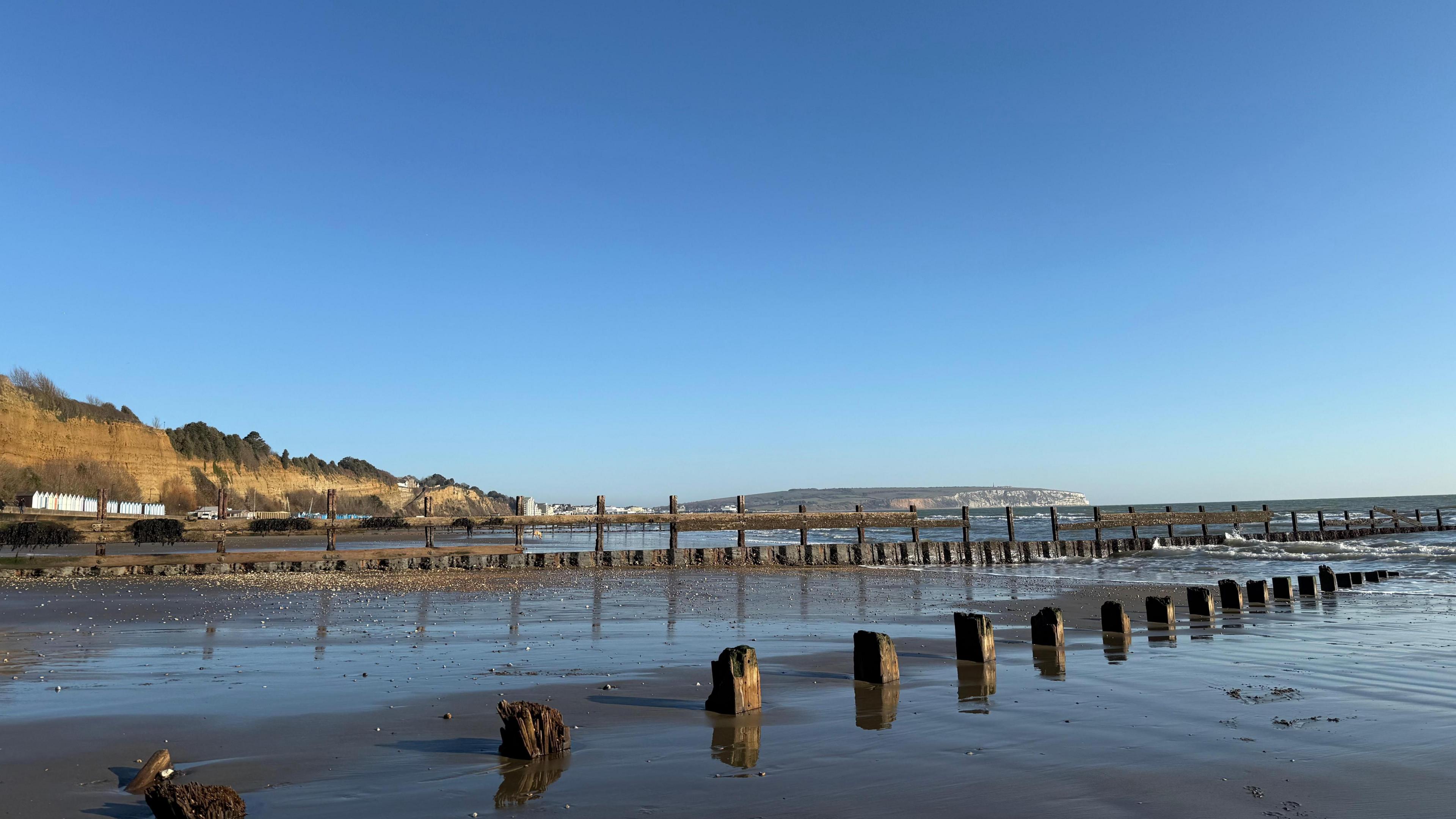 A sandy beach with wooden groynes poking through the sand. The sea is out and you can see small waves on the shore. In the distance you can see grass-topped cliffs. 