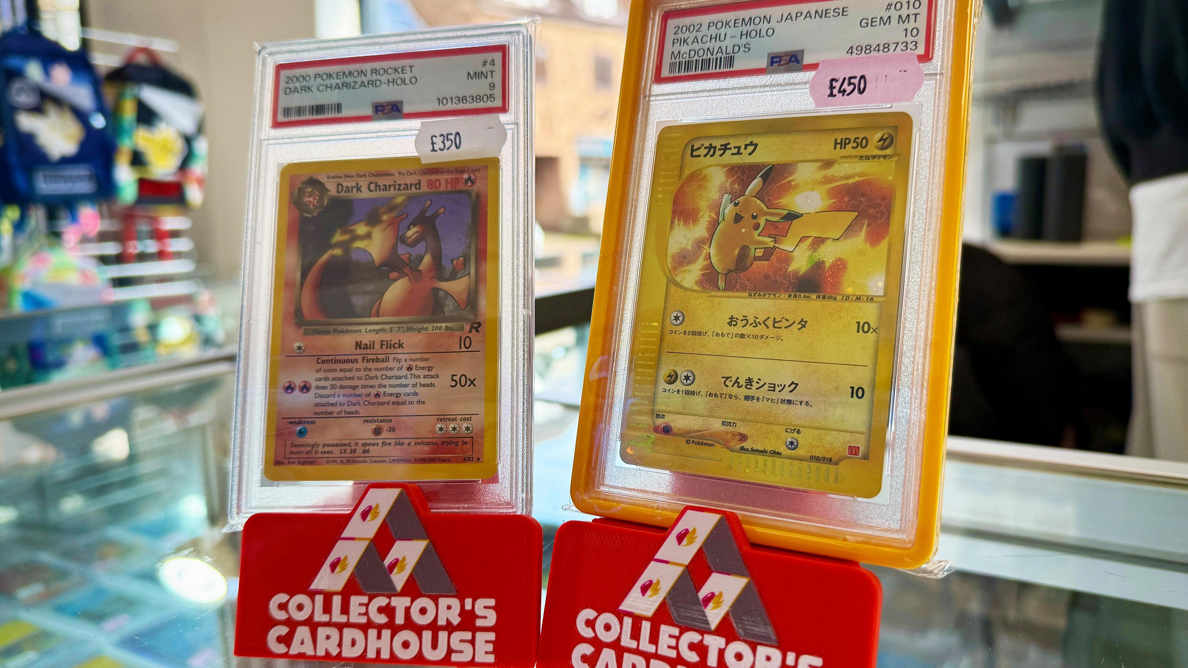 Two Pokemon cards stood upright on a counter in Collector's Cardhouse branded displays. One is a Dark Charizard card, the other is Pikachu card with Japanese text. 