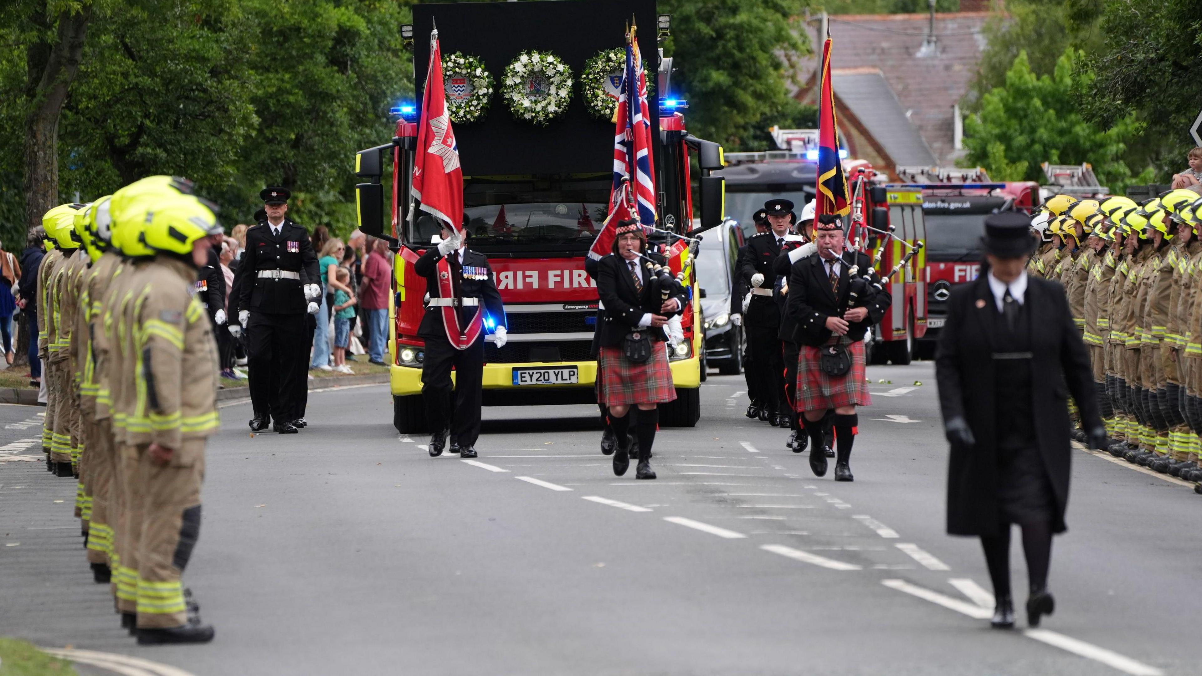 A view of the front of the funeral cortege of three fire engines, led by pipers in kilts and lined by firefighters in operational uniforms and yellow helmets.