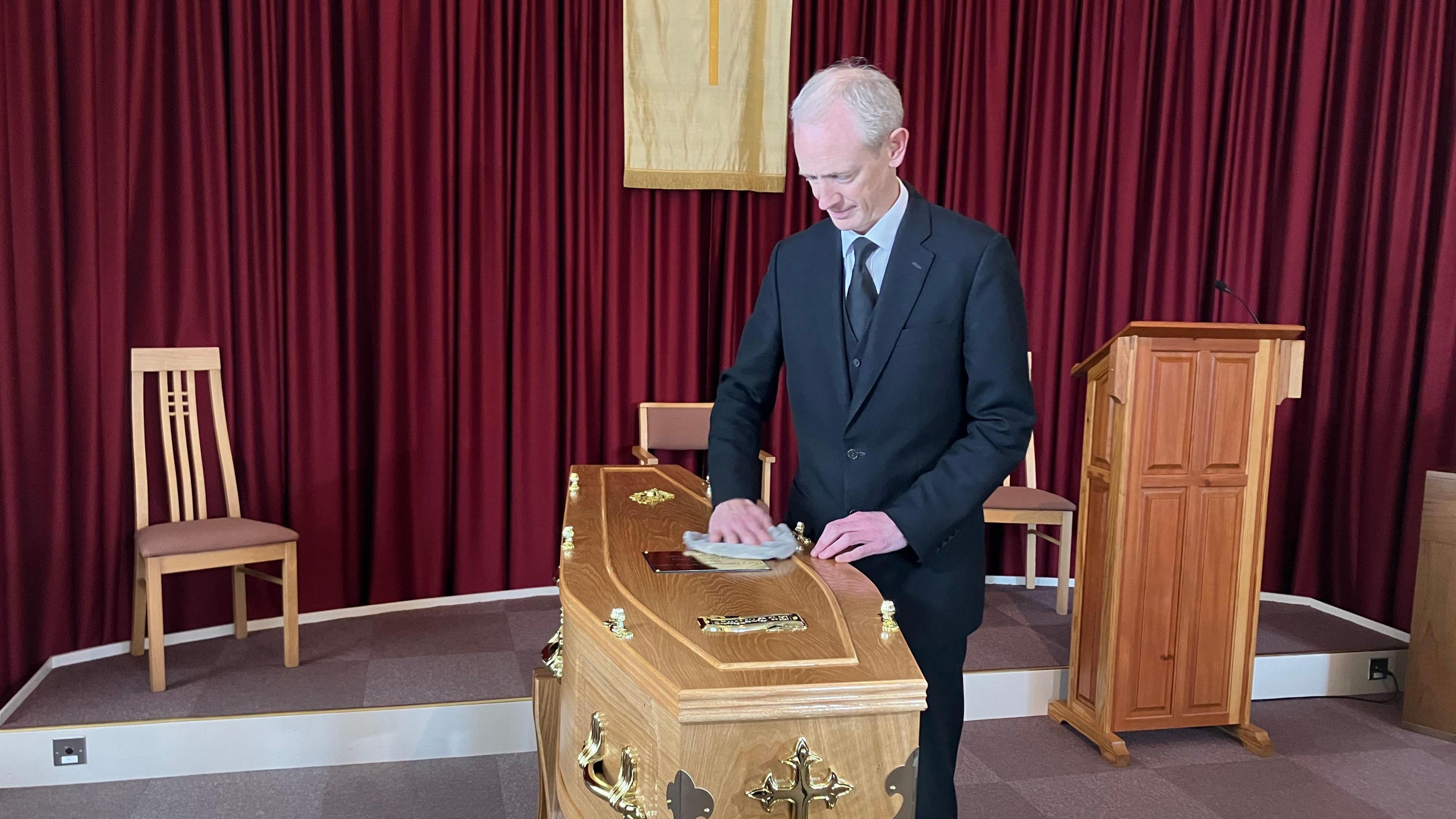 A grey-haired man in a black three-piece suit cleaning a coffin in front of a red curtain