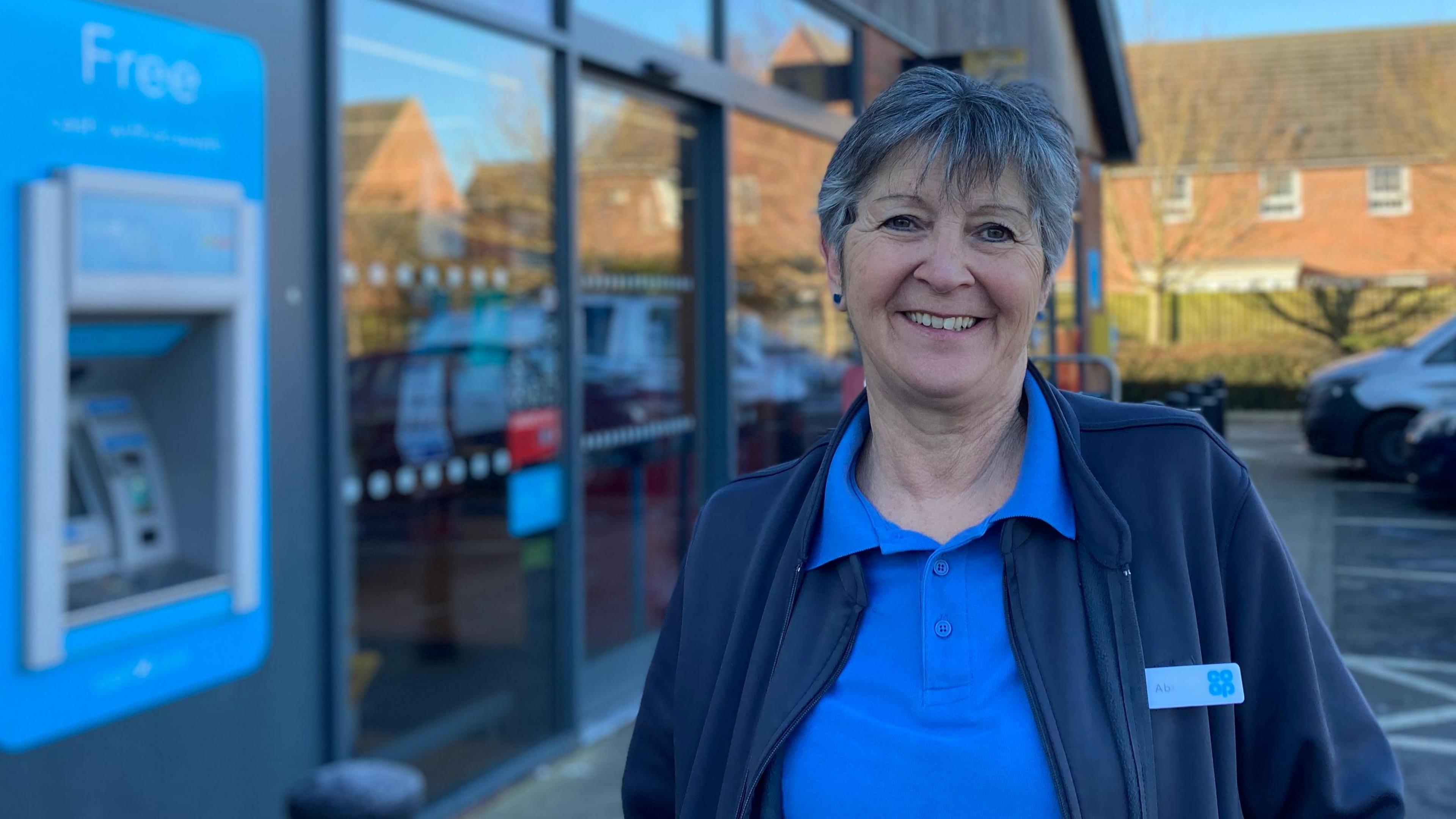 Co-op worker Abi Hill stands outside the glass store entrance. She has short dark hair and is smiling. She wears a Co-op blue polo shirt and fleece with a name badge.