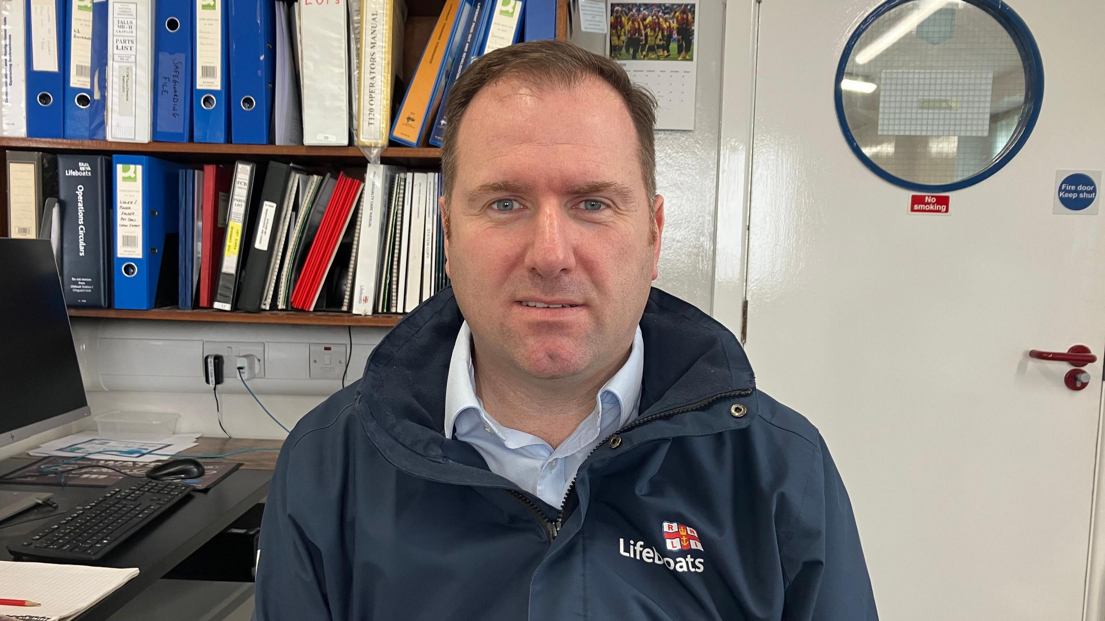 Daniel is looking at the camera and smiling. He is wearing a navy coat with 'Lifeboats' emblem on the left hand side. His shirt is white, his hair is brown and short and his eyes are blue. He is sitting in an office with folders behind him and a laptop.