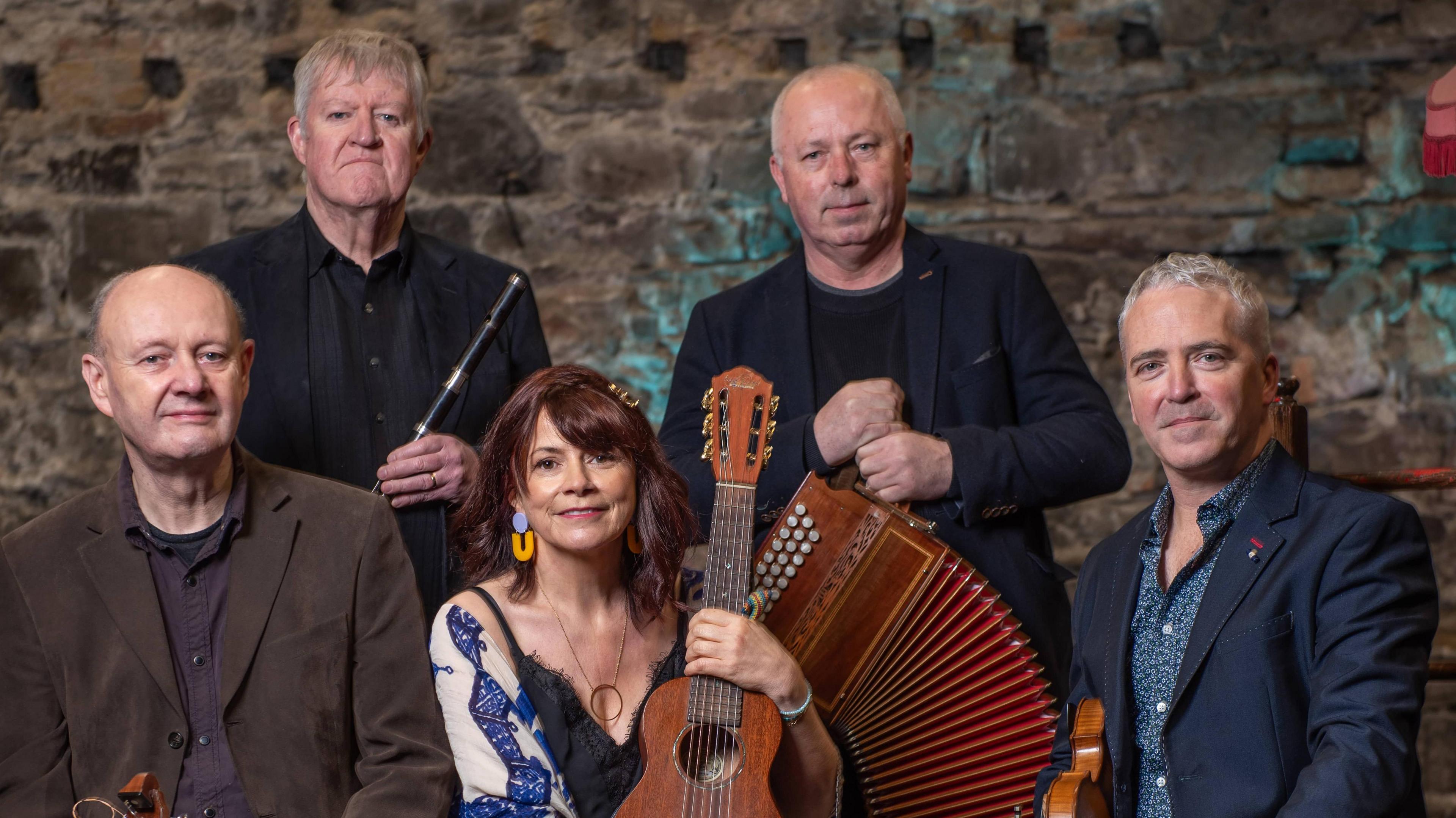 A group of older looking musicians.
Left to right: a bald man in a brown jacket sitting, a grey haired man in a black jacket holding a black whistle, a woman with reddish brown hair, blue and yellow earrings and a blue shawl over a black vest top holding a brown ukulele, a bald man in a black jacket standing holding an accordion and a man sitting with grey short hair in a navy jacket with a brown fiddle under his arm.