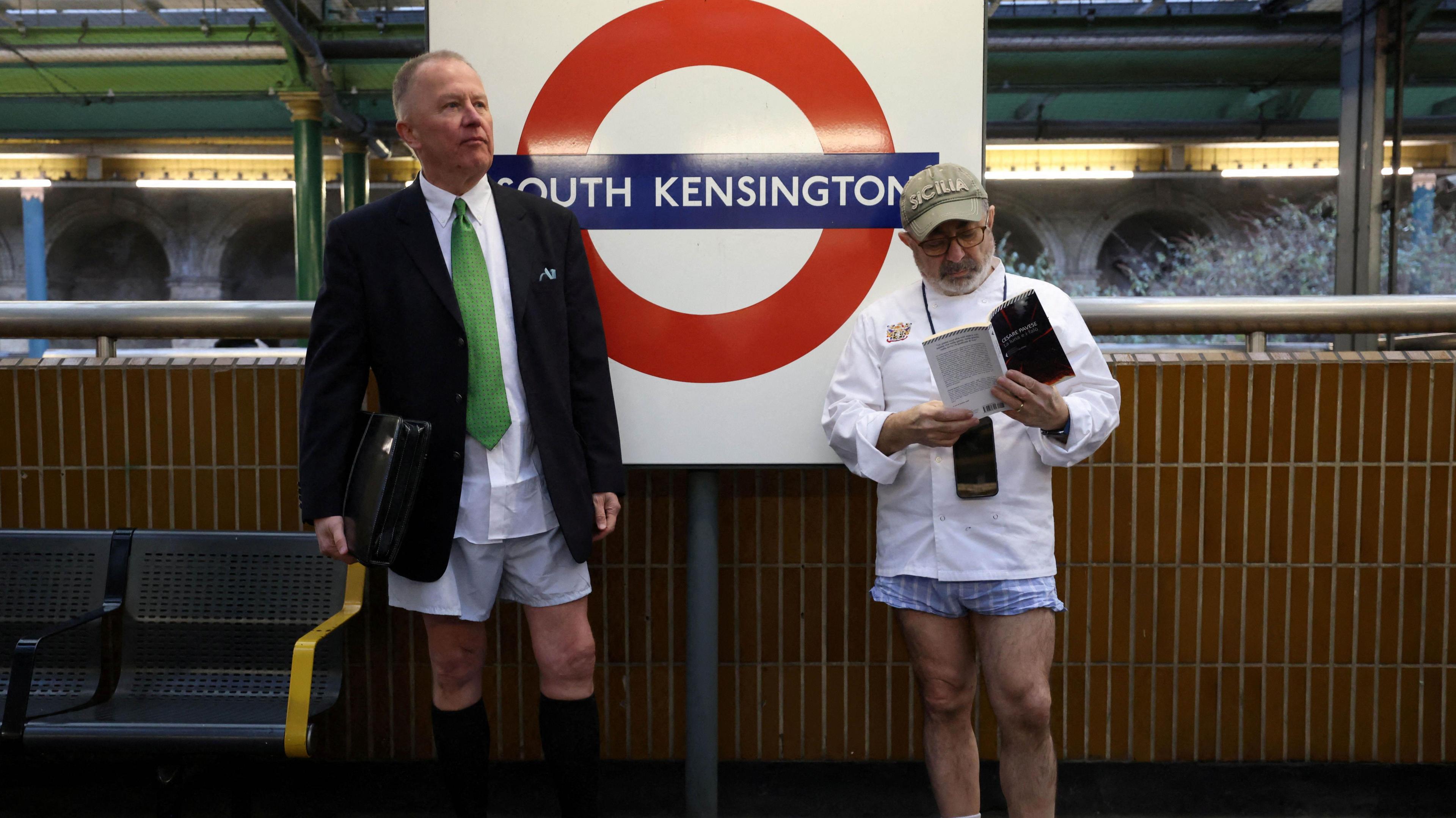 Two men without trousers, one reading a book, the other in a suit and tie in front of South Kensington Tube roundel