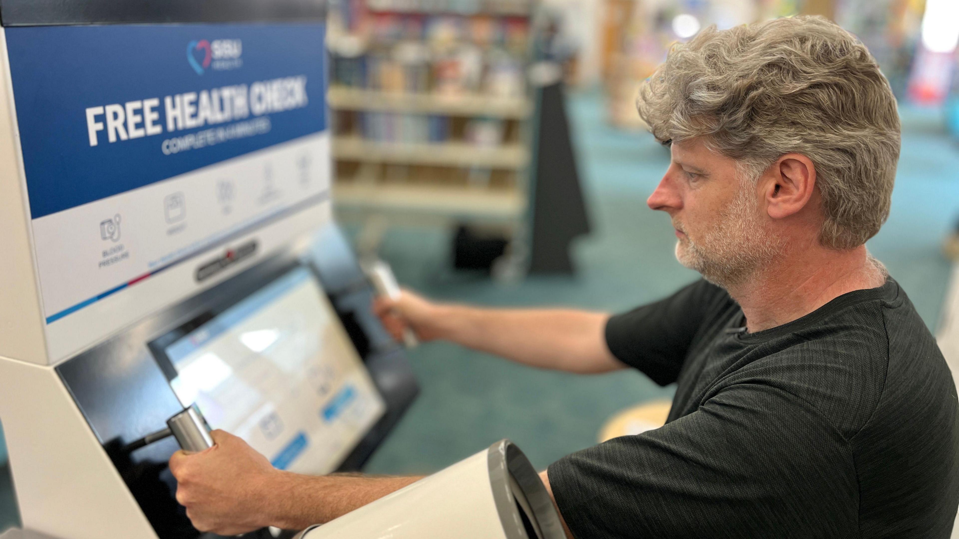 Michael Hadley puts his hands on two metal handles which use electric pulses to measure body fat. He is looking at the screen which guides him through the process in the corner of the library