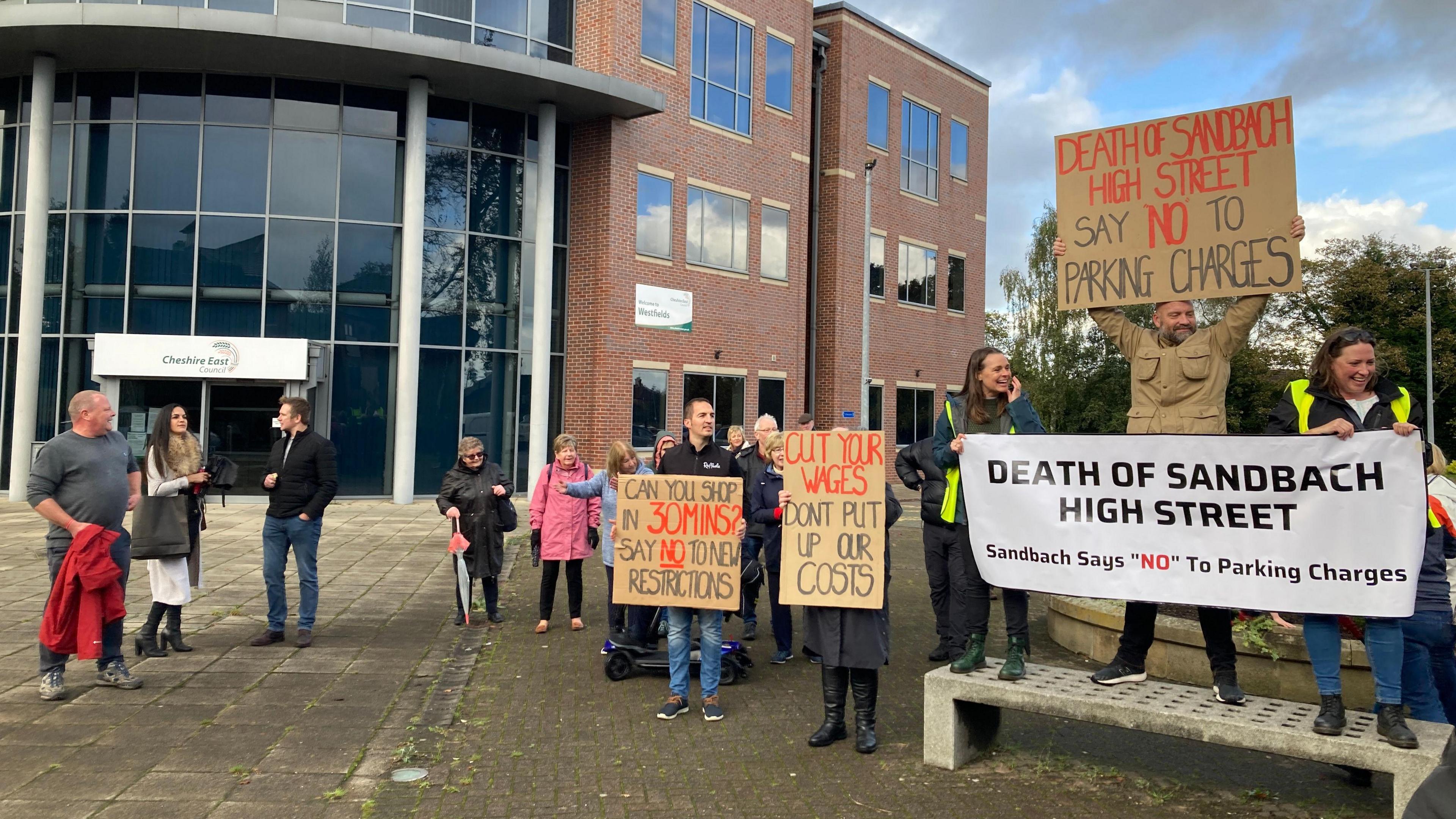 Protestors from Sandbach outside Cheshire East Council's headquarters. They hold cardboard signs in red and black letters with phrases including "Can you shop in 30 mins? Say no to new restrictions" and "Cut your wages, don't put up our costs". They also have a white banner with black letterings saying "Death of Sandbach High Street. Sandbach says "no" to parking charges". The protestors wear coats and stand in front of a red-brick building with a glass atrium which has a "Cheshire East Council" sign on it. 