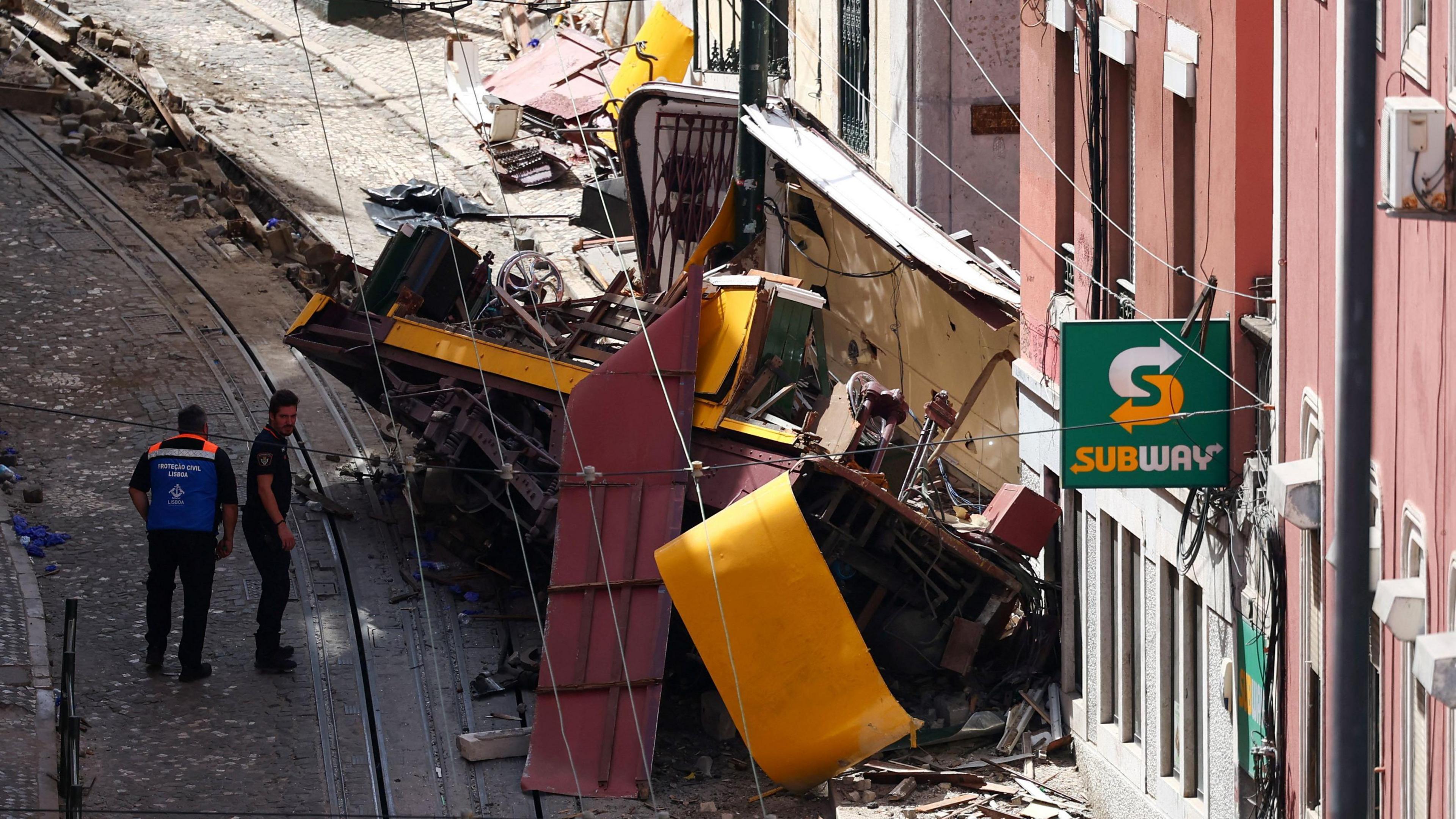 The crumpled metal yellow and white funicular is seen on its side against a building in Lisbon, beside a Subway restaurant sign, as two male emergency workers walk on the cobblestoned street.