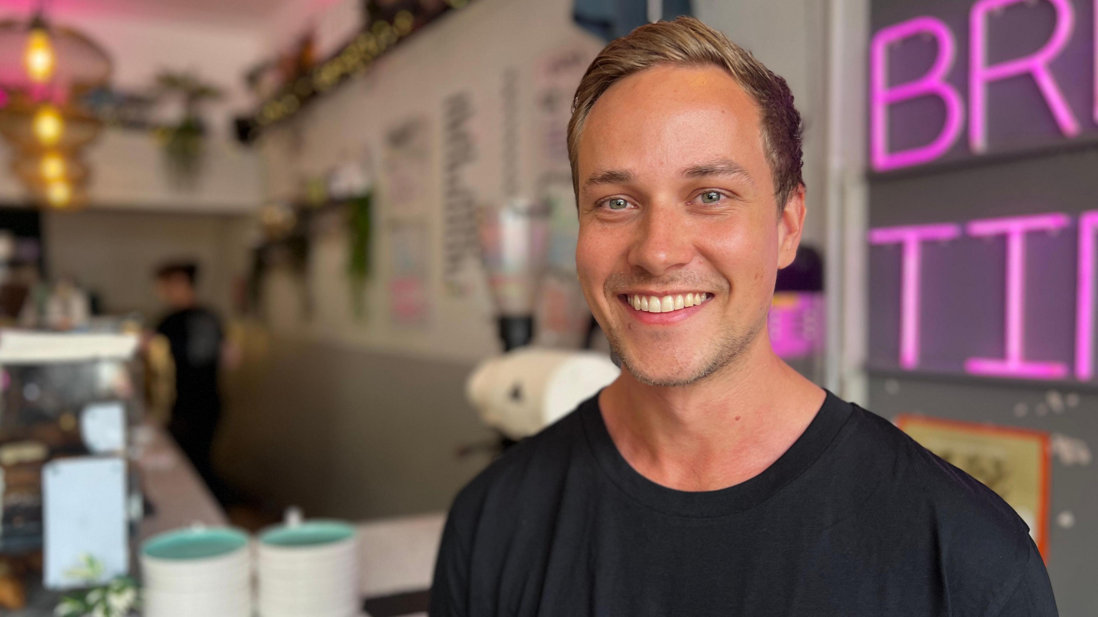Cary Frame, owner of Corner Coffee Co in Cardiff, smiles at the camera. The coffee shop is in the background. He has mousy blonde hair and a black t-shirt on.