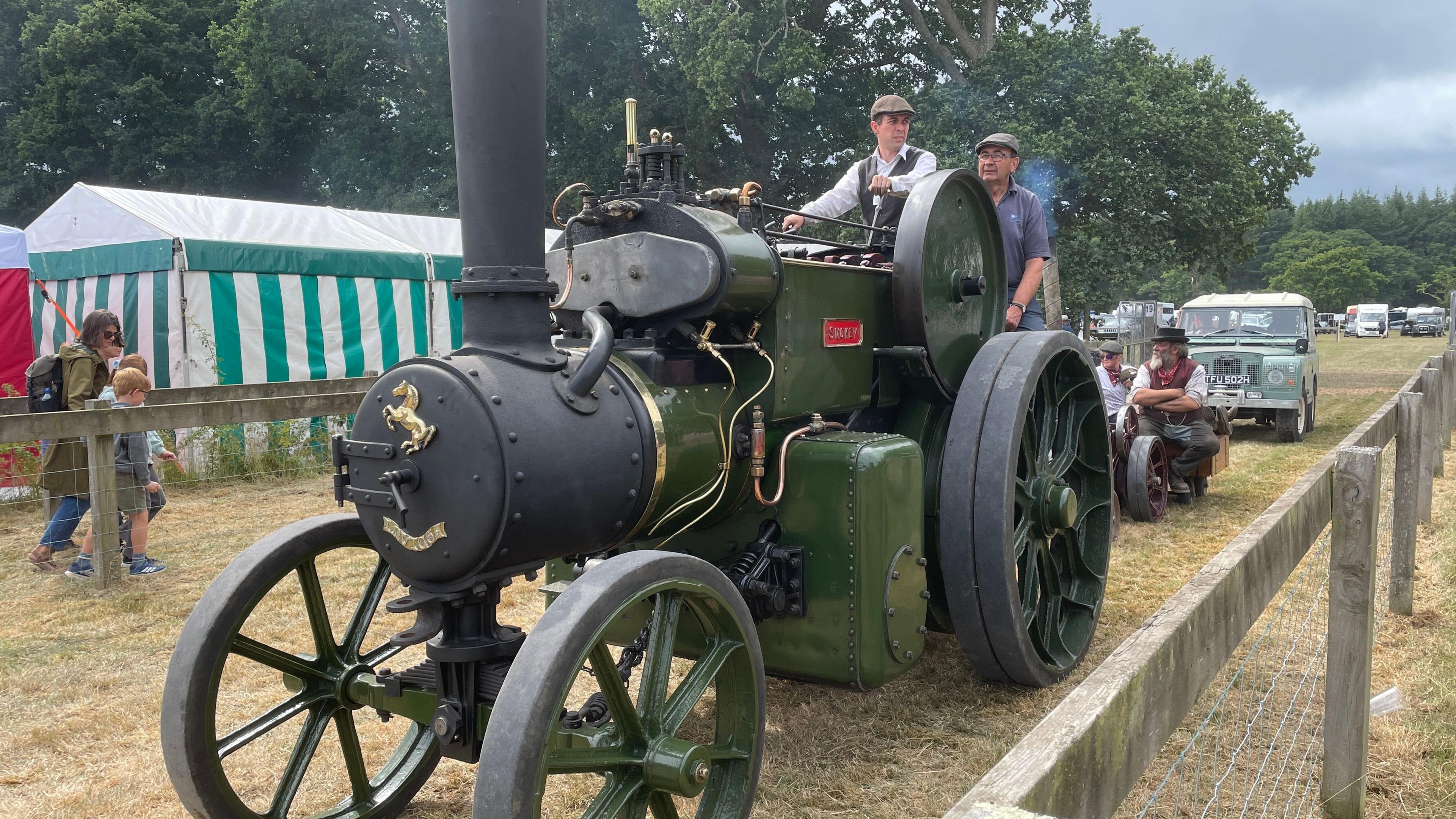 A green retro tractor operated by two men in flat caps. It is in a field with other retro vehicles in the background. 