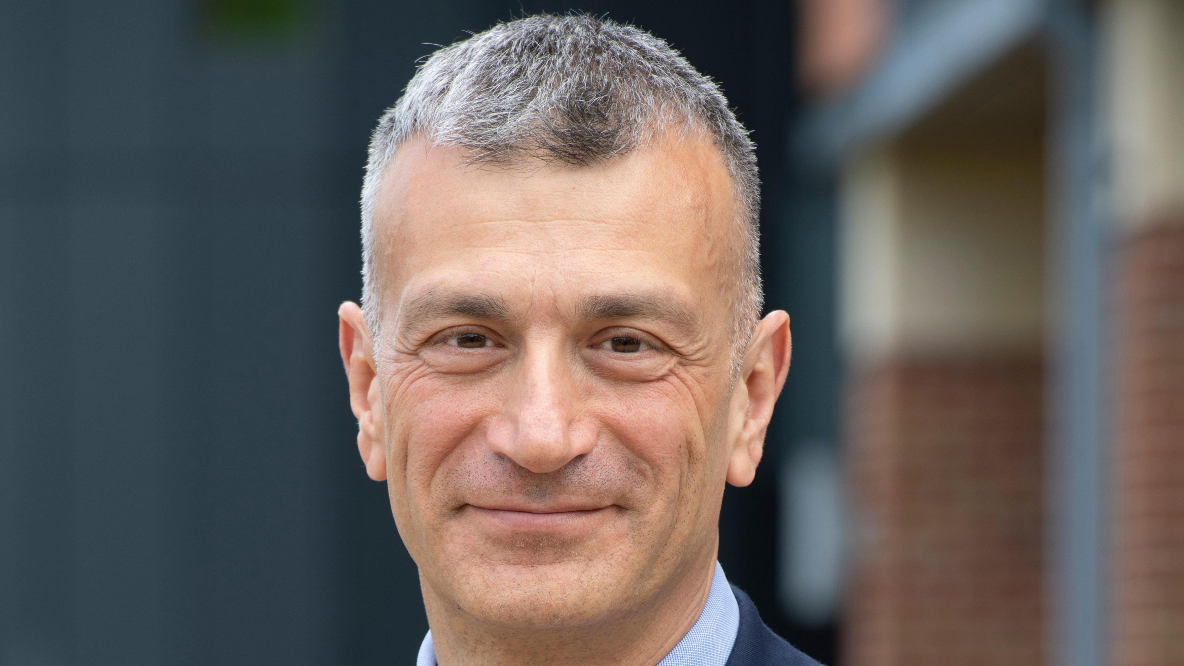 A man with black and grey short hair smiles at the camera. He is wearing a navy suit with a light blue shirt underneath. The background behind him is blurred.