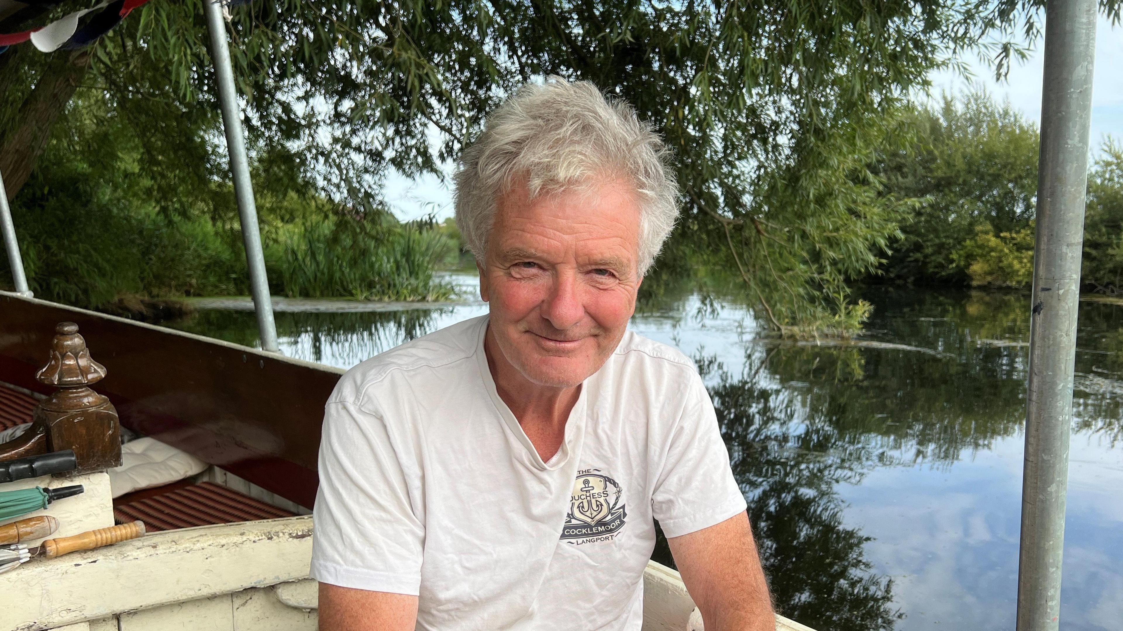 A man who has grey hair is looking at the camera. He is sitting on a boat which is floating on a river. There is a tree dipping into the water, and lots of green around the riverbank.