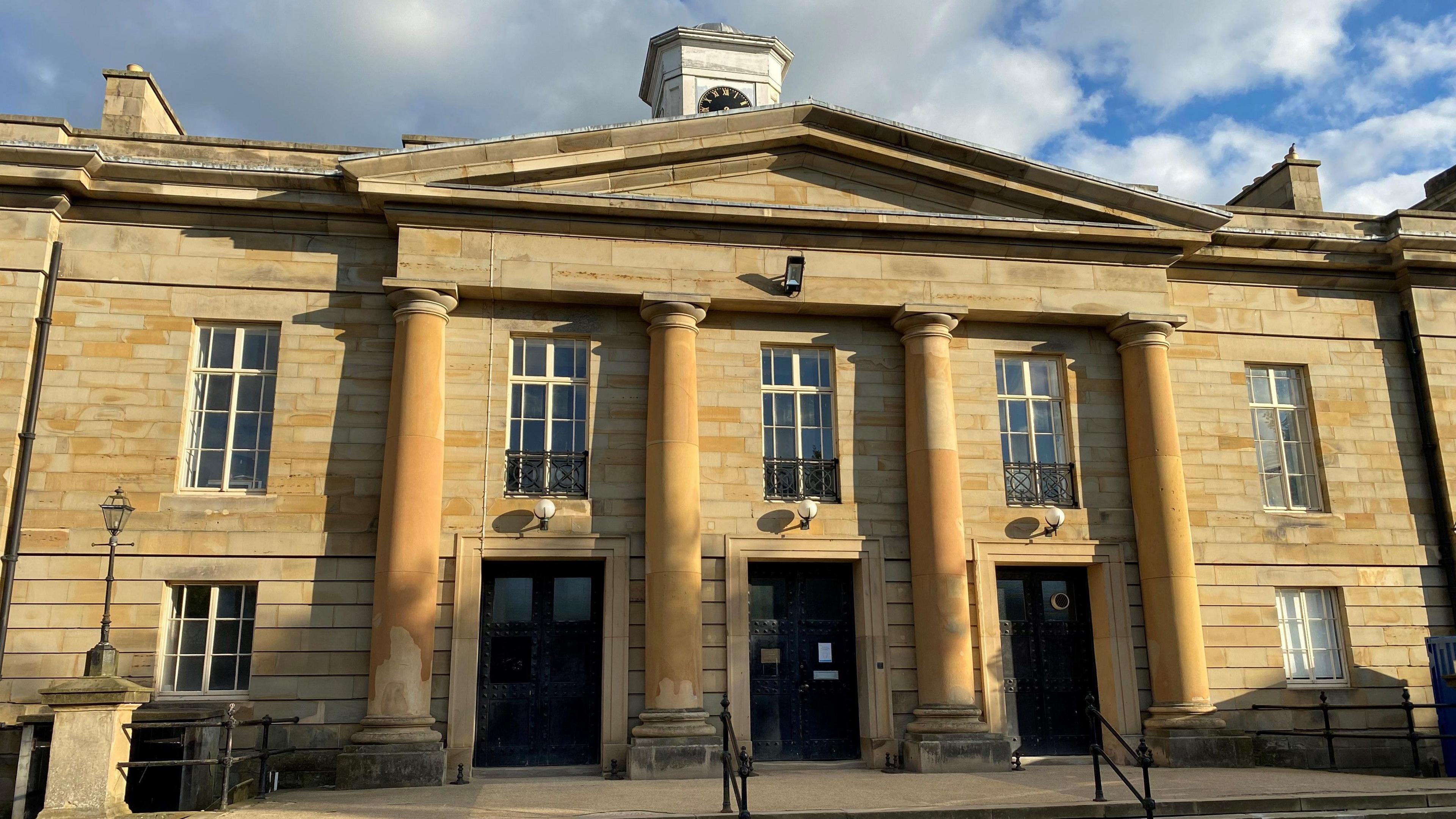 Durham Crown Court. It is a two-storey Georgian-style building with yellow stonework, large rectangular windows and three columns around the three large black doors.