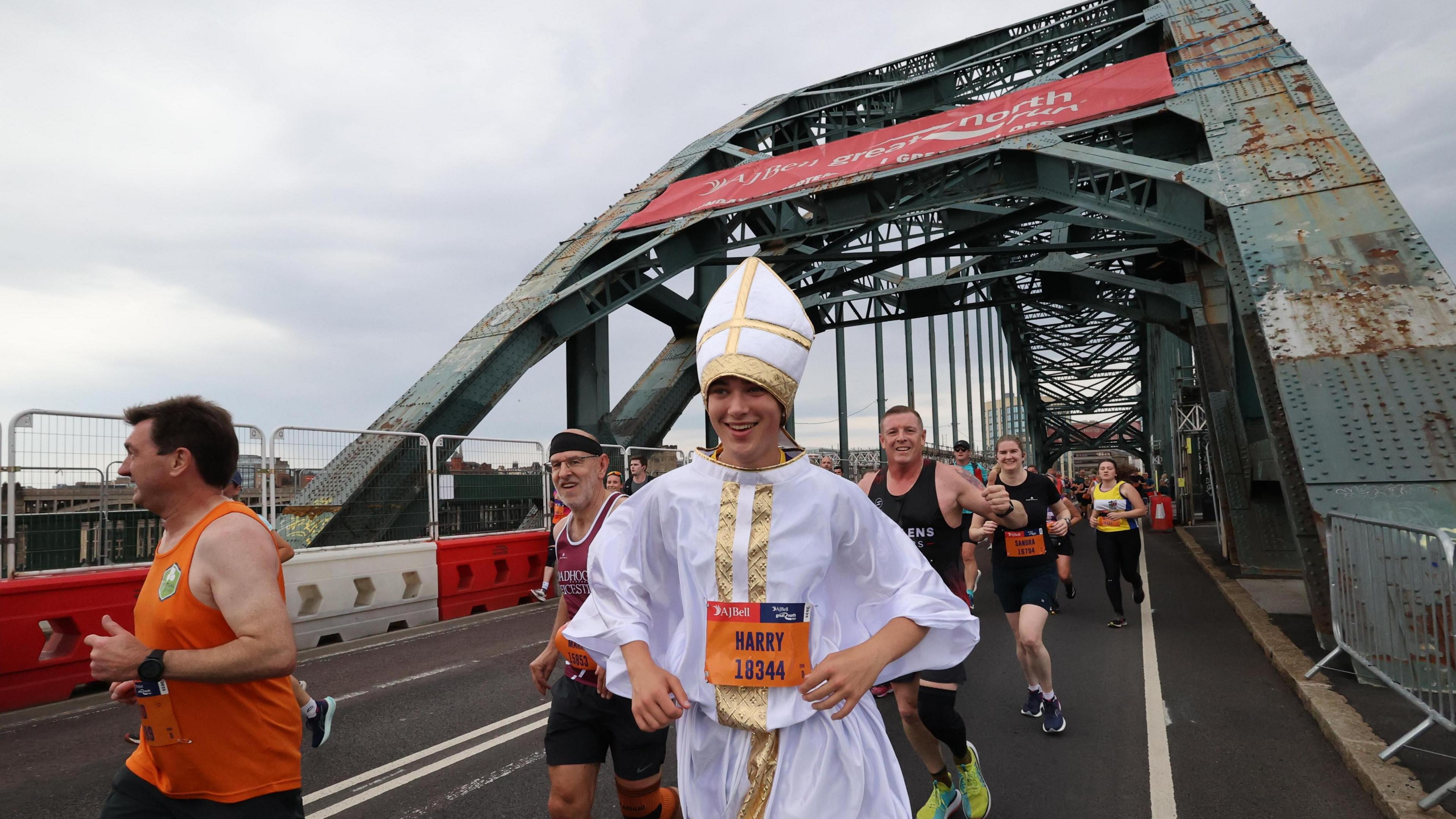 Young man dressed in pope costume runs across the Tyne Bridge. There are several runners on the bridge nearby. Everyone is smiling.