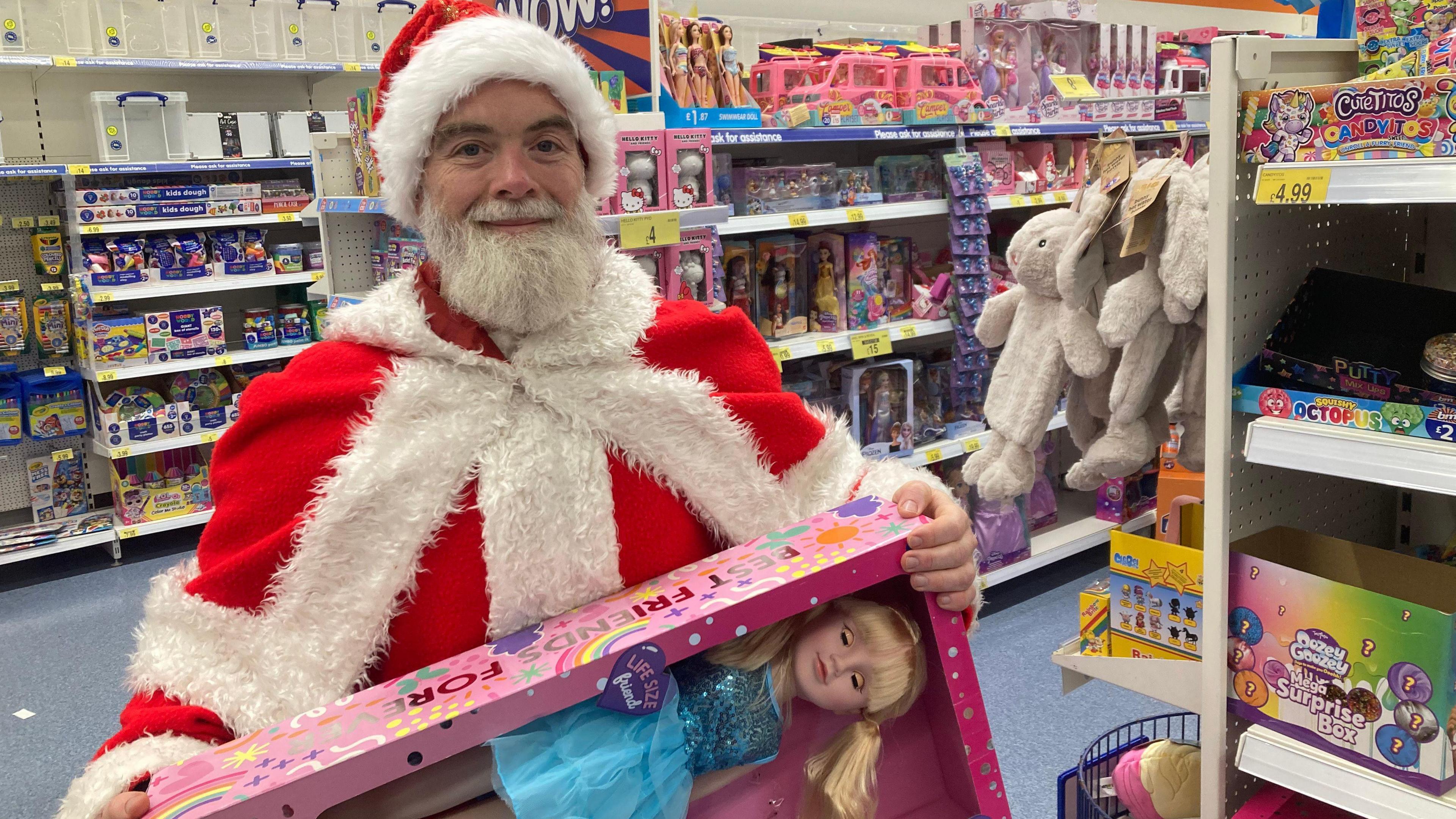 A bearded man stands wearing a red and white Santa costume. He is standing inside a toy store and holding a blonde doll in a blue dress which is in its box. 