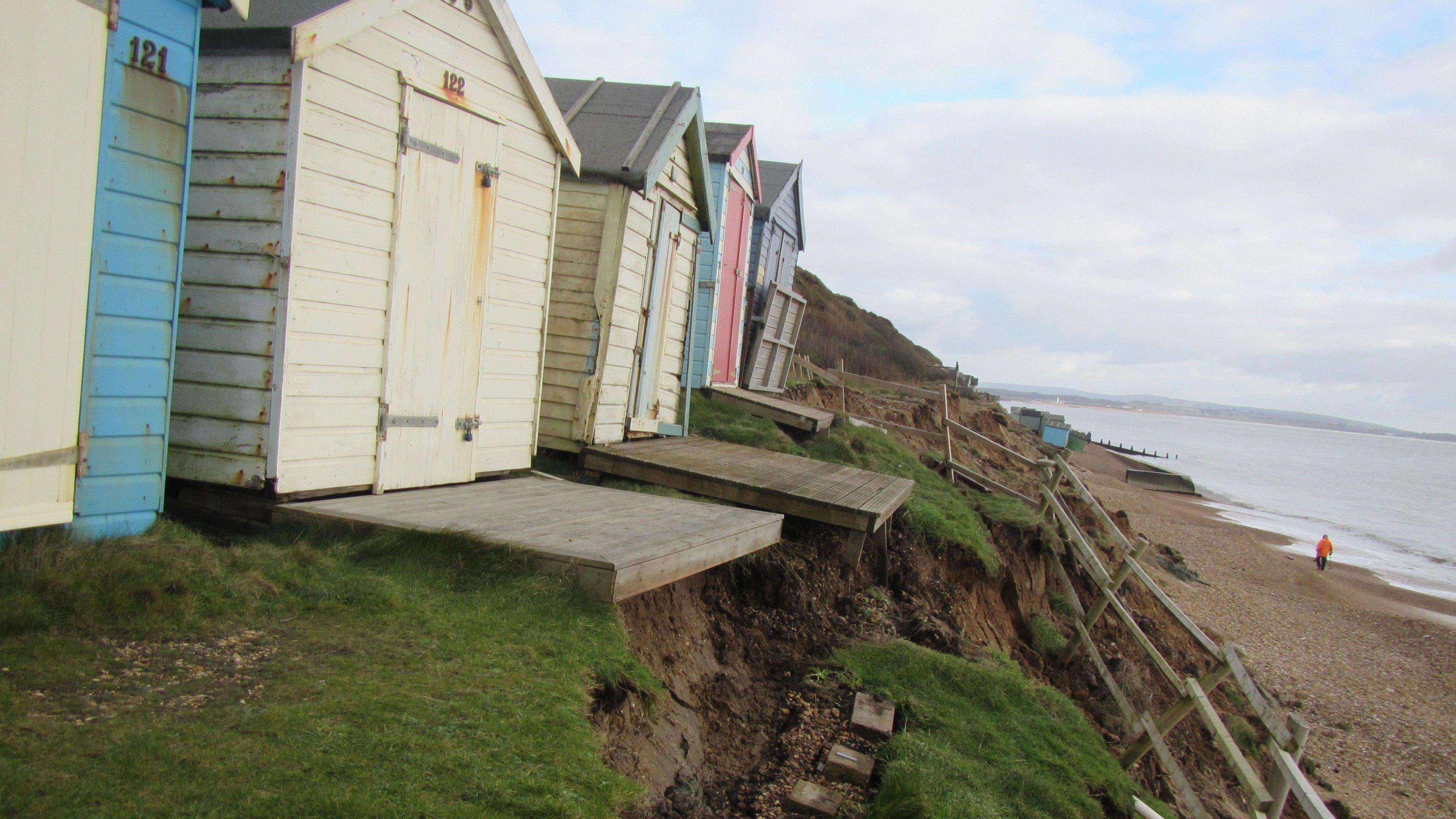 Five beach huts on the edge of a cliff leading down to the beach. Some of the huts are leaning close to the edge