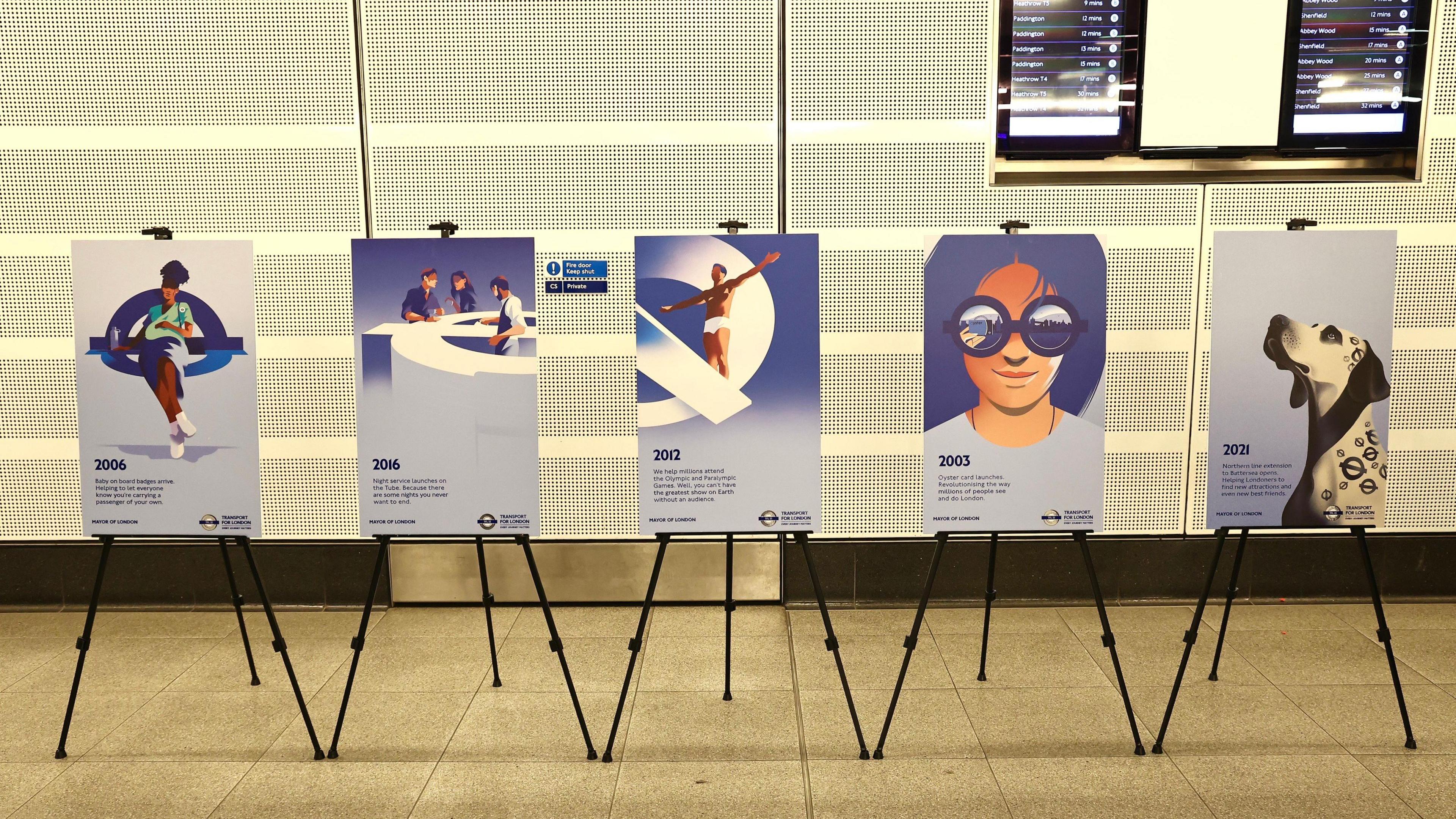 Five blue-toned artistic posters show dates and descriptions of events from TfL's history. They are displayed on black easels in front of a white wall inside a TfL station