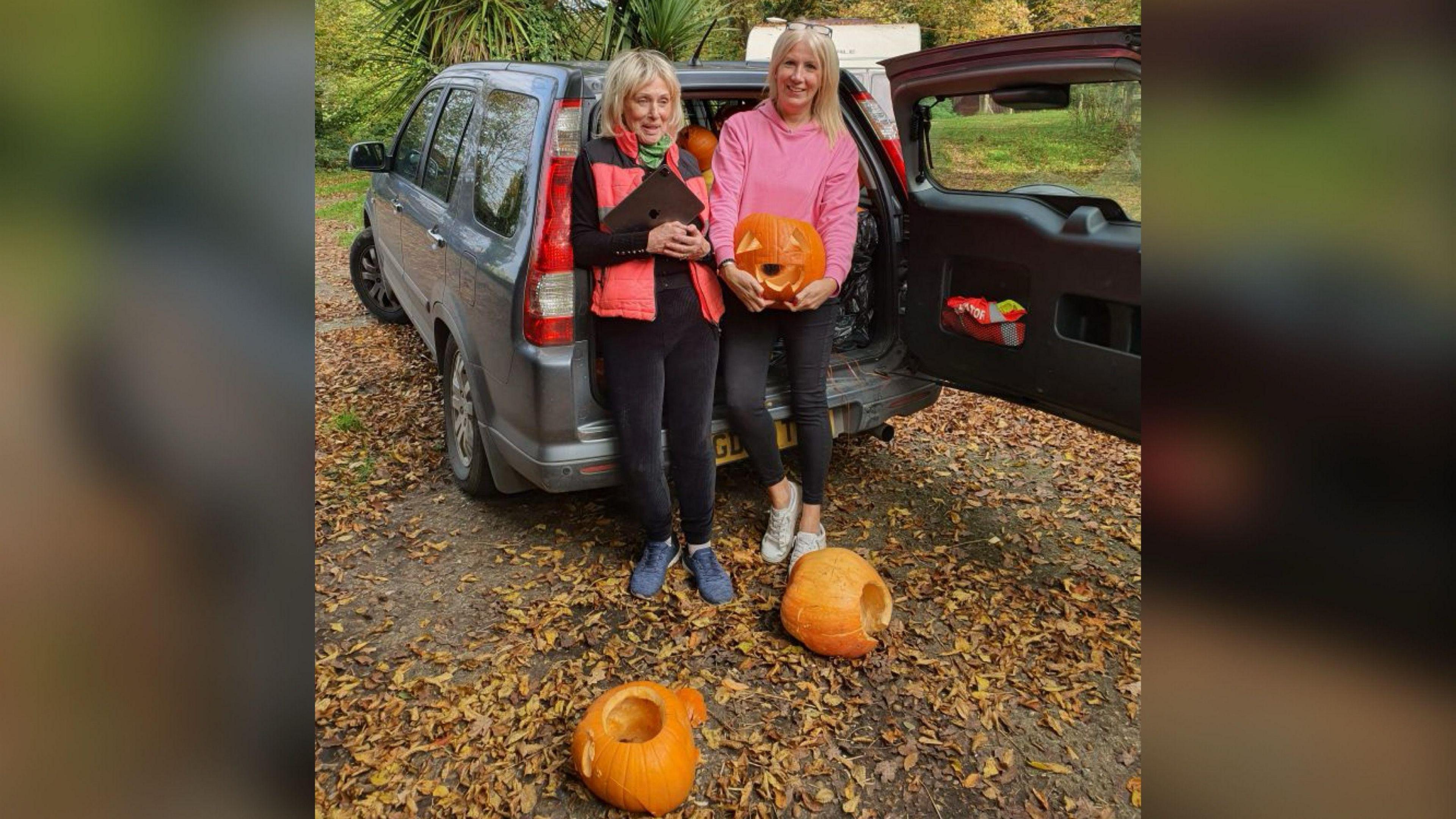 Two blonde women in pink coats leaning on a car with its boot open. One of them is holding a carved pumpkin while two others lay on the ground in front of them.