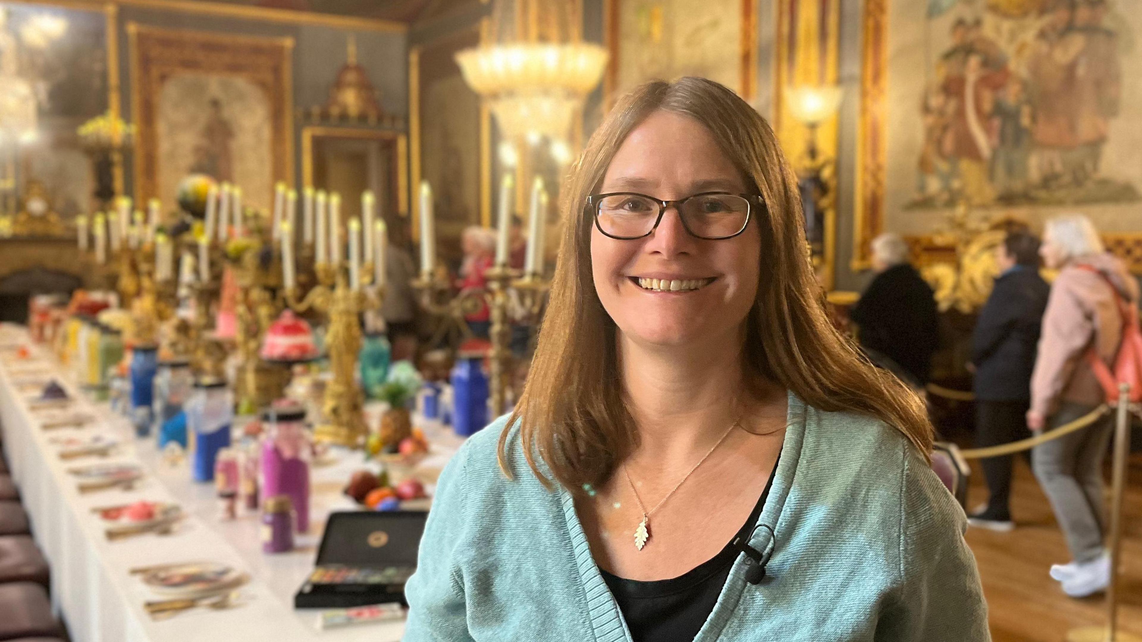 A smiling woman with glasses, wearing a black dress and light blue cardigan, is looking into the camera. Behind her is a lavish dining table with candelabra and colourful place settings. There are visitors in the background of the dining room, which has chandeliers and ornate floor-to-ceiling paintings.