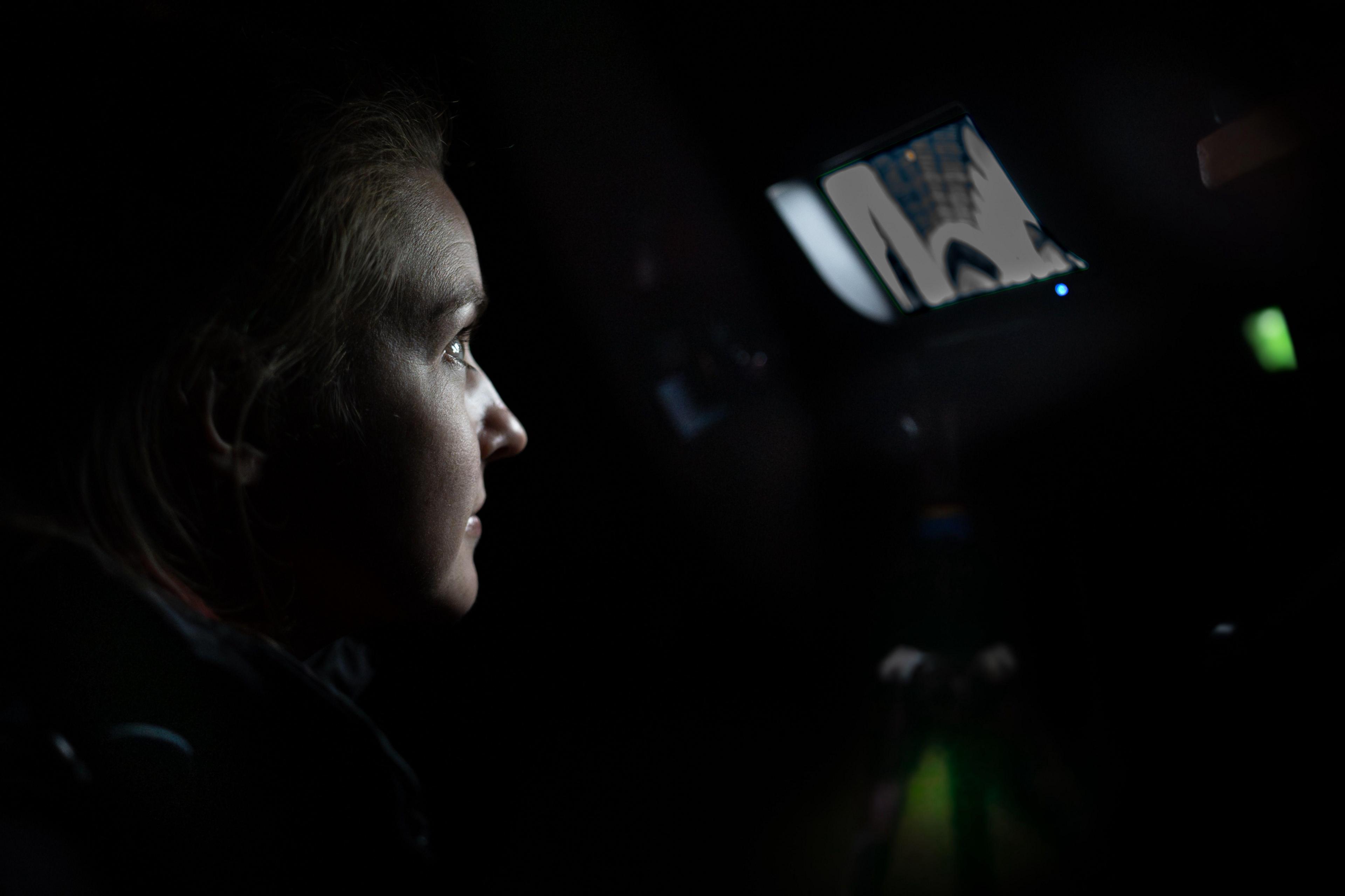 A woman looks into a screen in a church at night. the image is mainly dark, apart from a small screen that shows an image of the roof of church. It glows in the darkness, illuminating the woman's face who is staring intently at it.