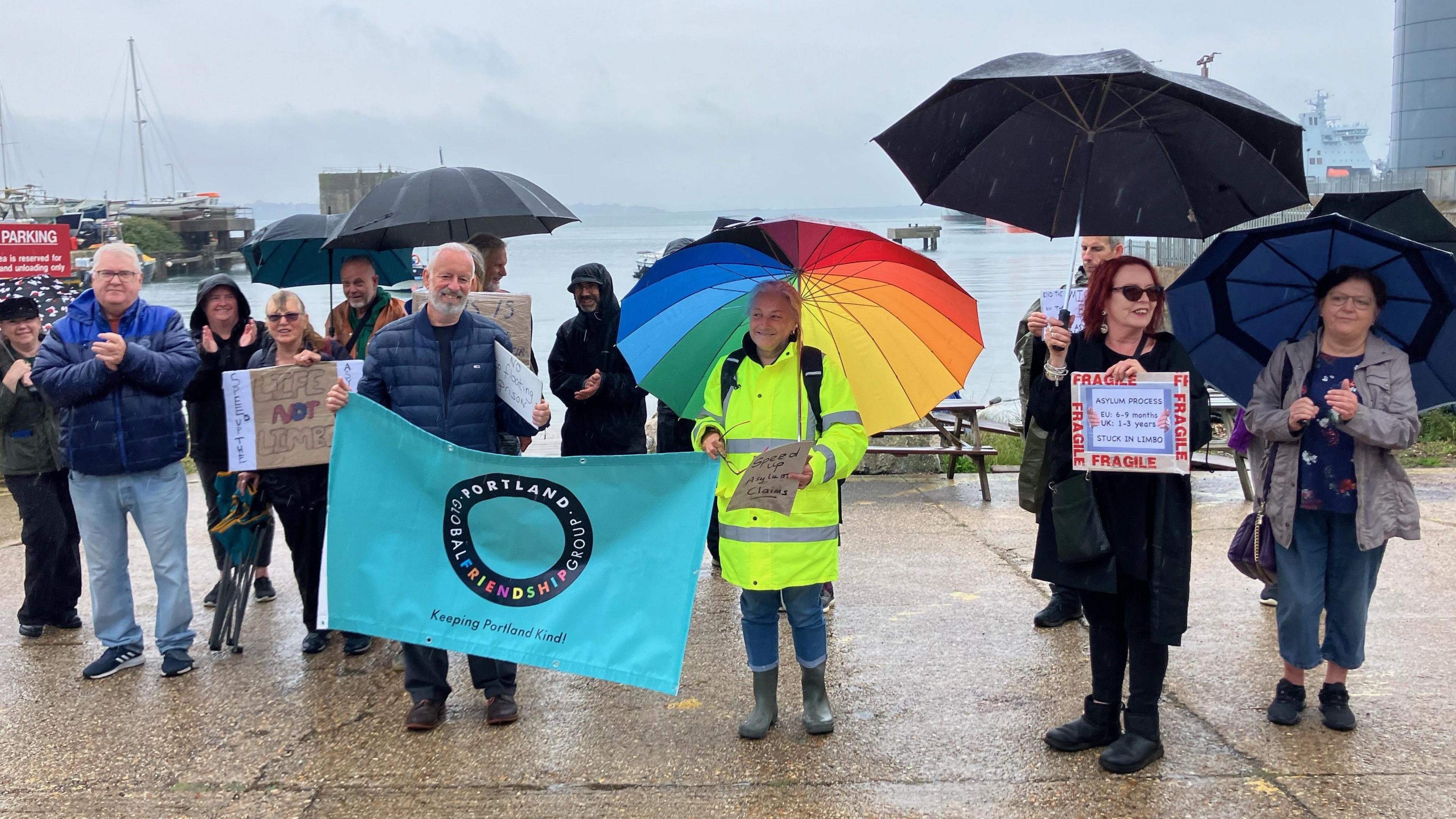 About a dozen protestors standing in the rain. They are holding placards and umbrellas. Behind them is the sea.