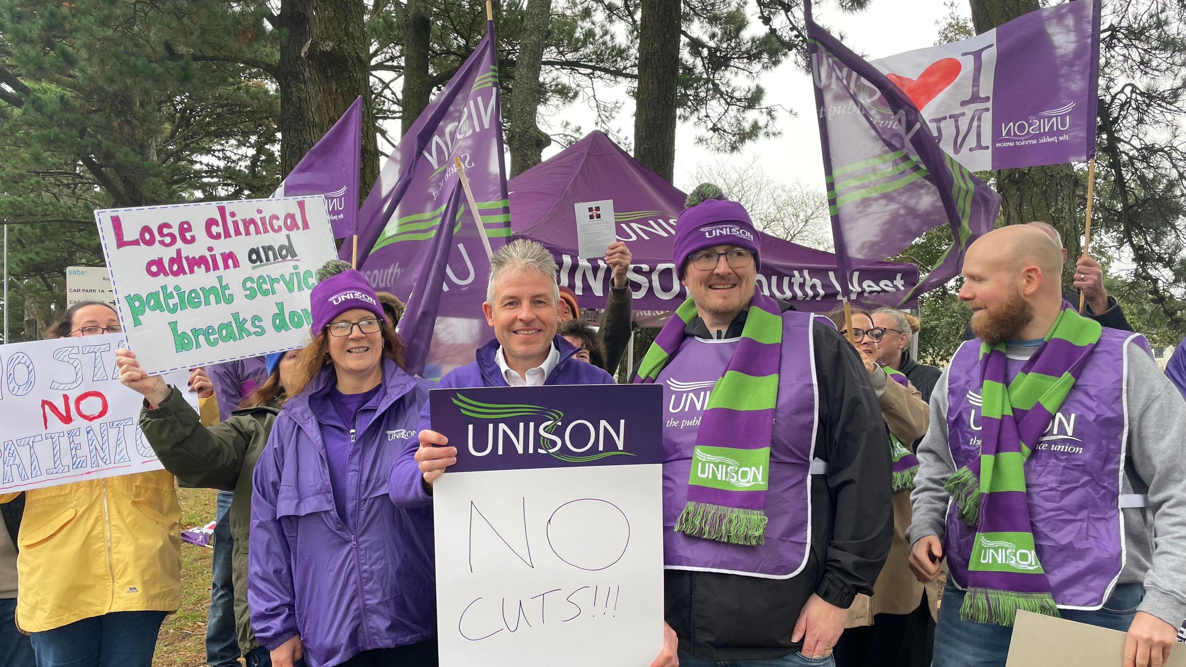 About nine people, in purple and green clothing, holding placards. Some people in the back are holding UNISON and "I LOVE NHS" flags. There are trees in the background. The sky is grey.