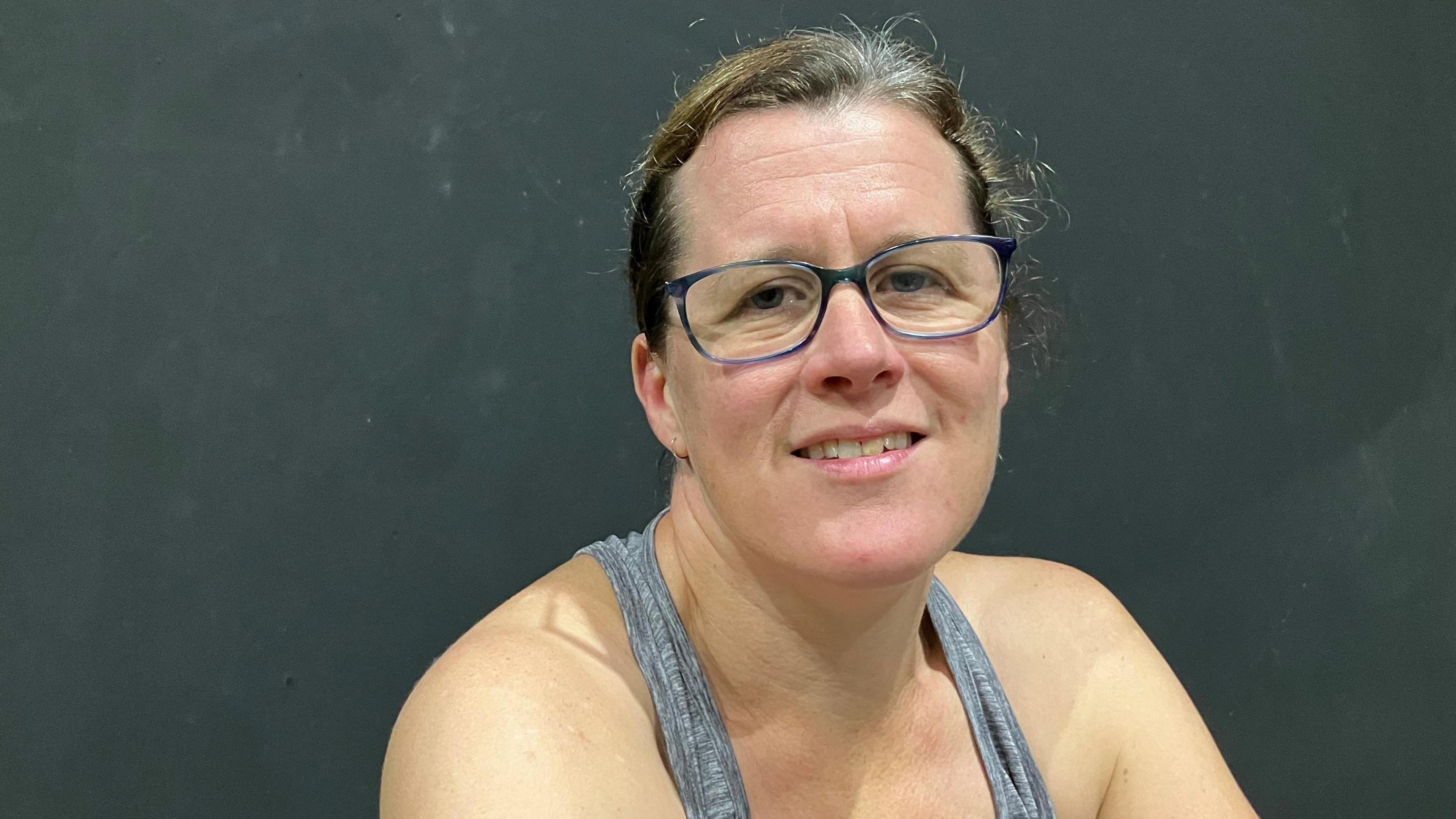 A woman with her hair tied back, wearing glasses and a grey vest in a gym with a black wall behind her. It is a head and shoulders shot.
