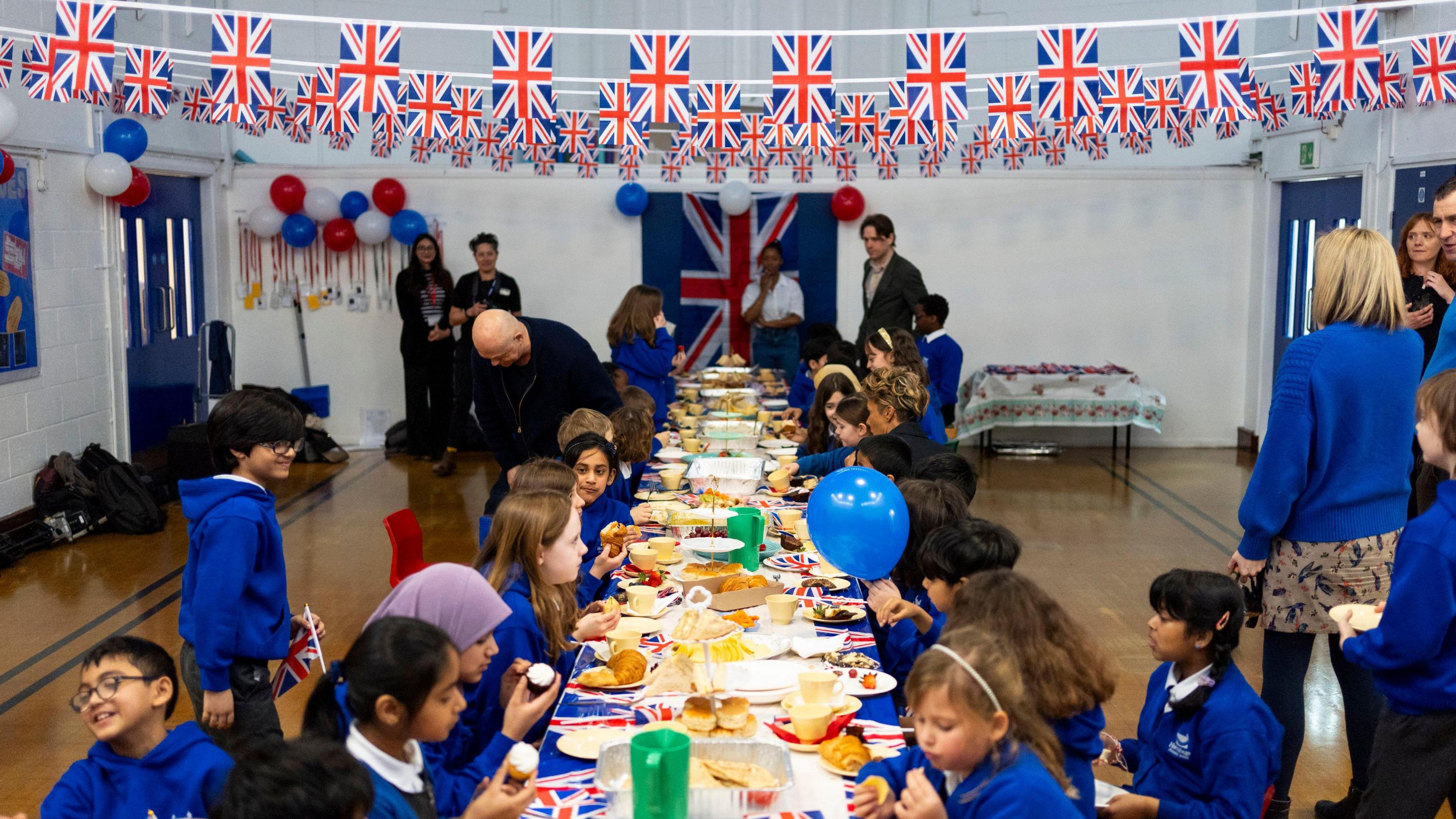 Children in school uniform sitting at a long table eating with union flag bunting hung from the ceiling 