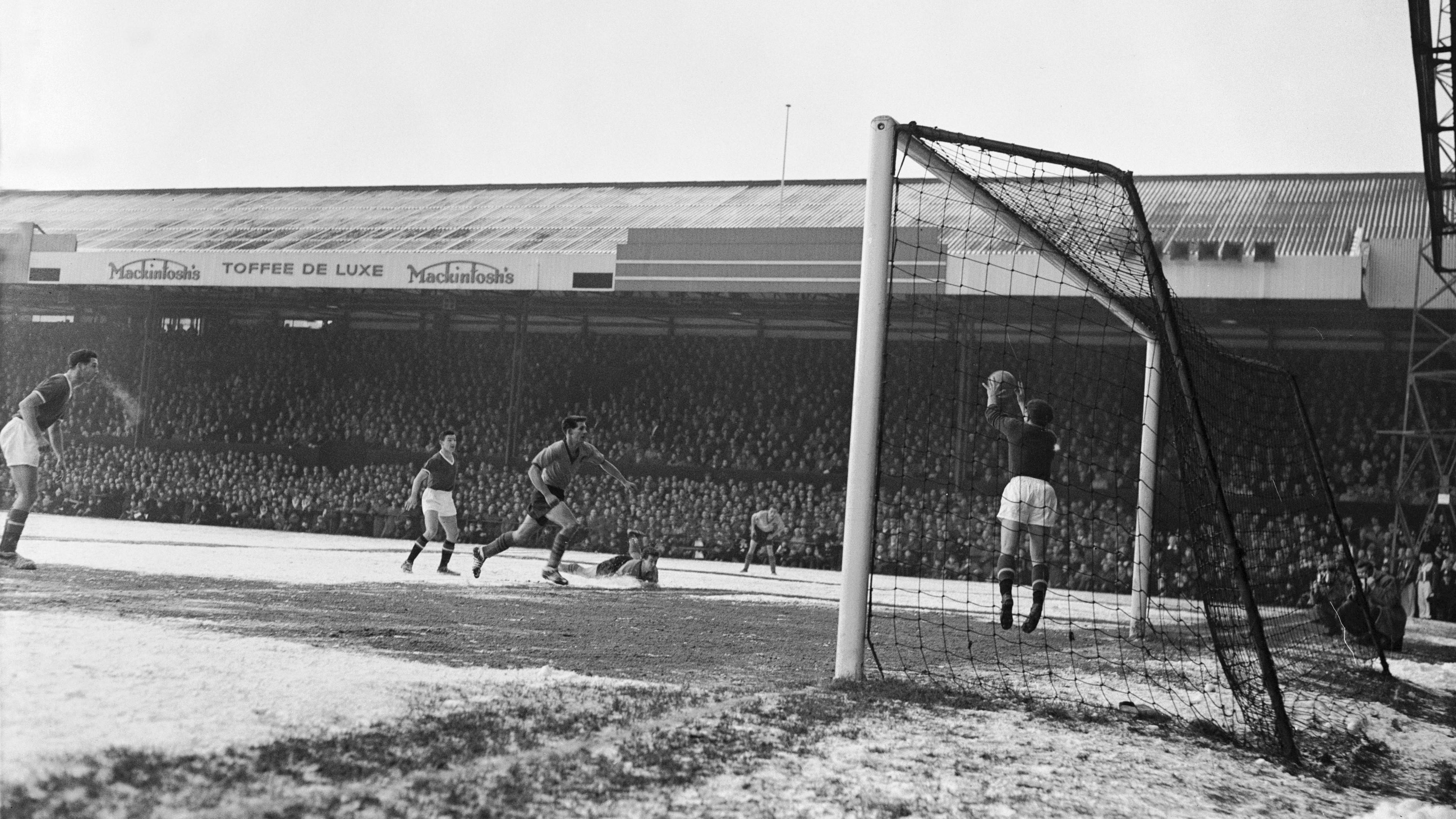Black and white photograph of Norwich’s football stadium during a 3–0 victory over Manchester United. The photo is taken next to goalposts with a view across the penalty box. The goalkeeper has the ball in his hands just above his face. Five other players are in the box with one on the ground and one appears to be running towards the goalkeeper. The roofed stand in the background runs along the far side of the pitch and appears to be full. There is frost on the ground.