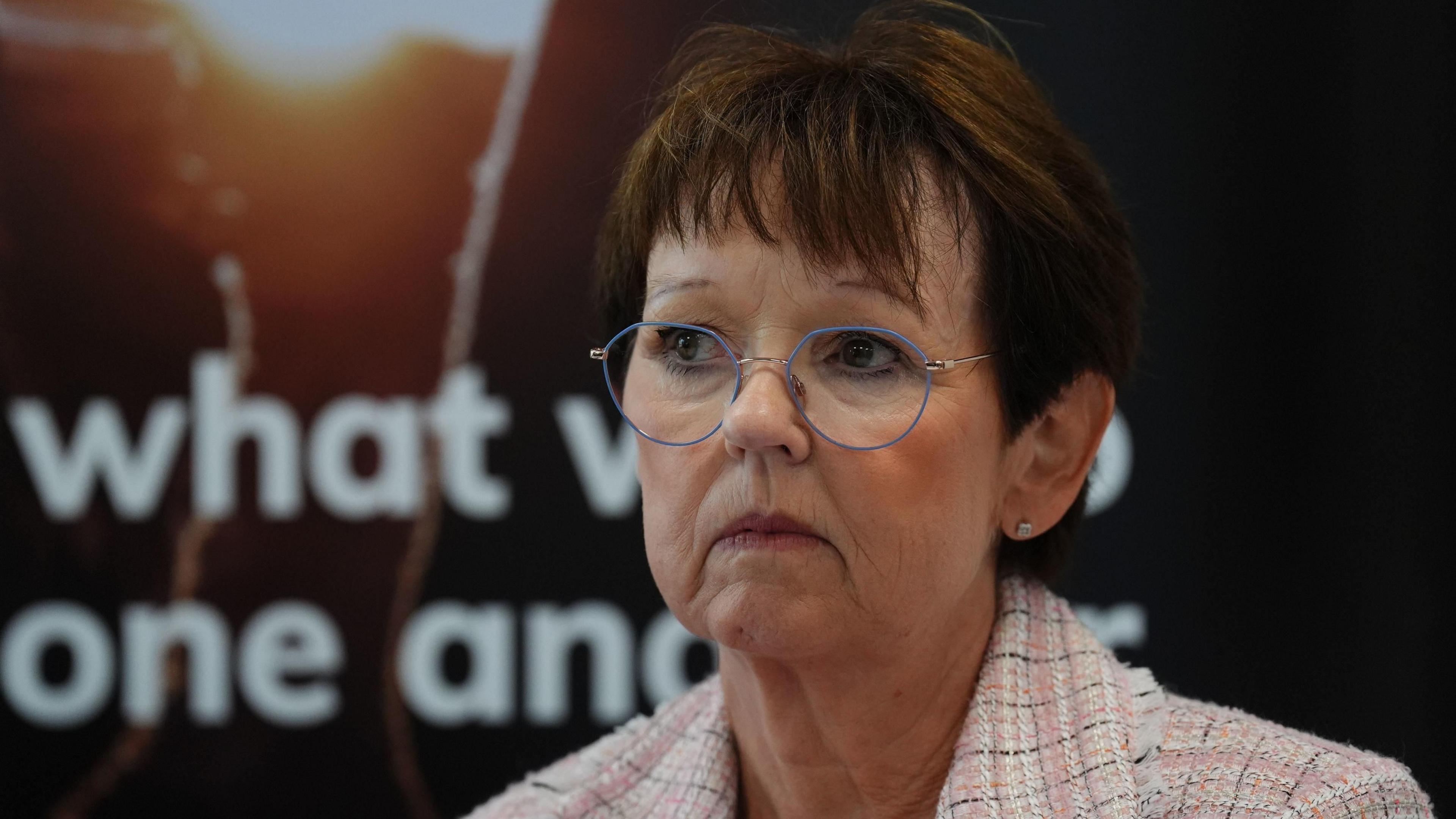 Carol Gillies at the press conference, sitting in front of a poster. She is wearing a pink checked jacket and glasses, and has short brown hair.