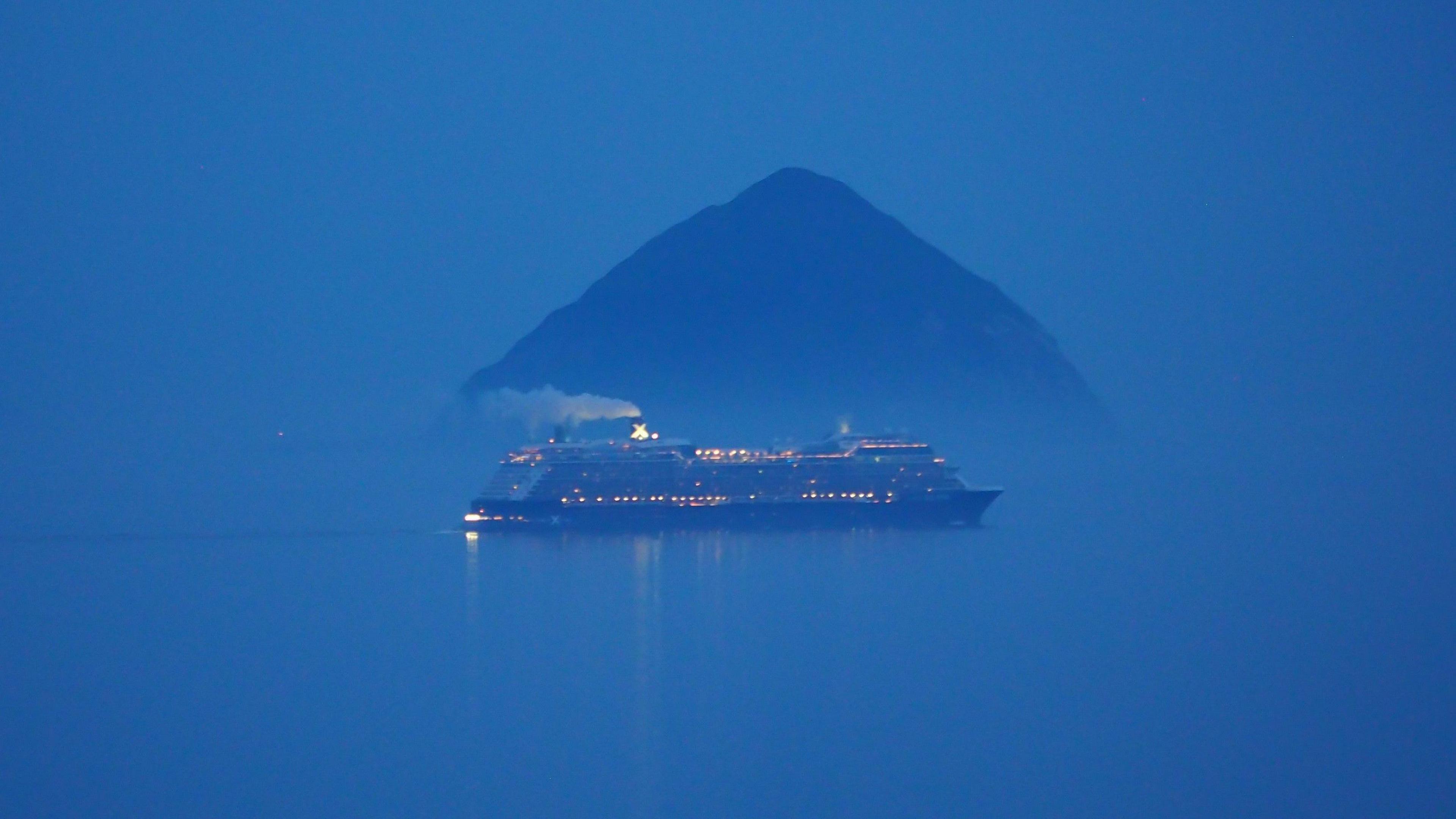 A cruise ship, with lots of lights and a large plum of smoke coming from the top sailing in front of Ailsa Craig, a large dark rock in the water. The ship and rock are the only thing visible as a dark blue sky and water merge.
