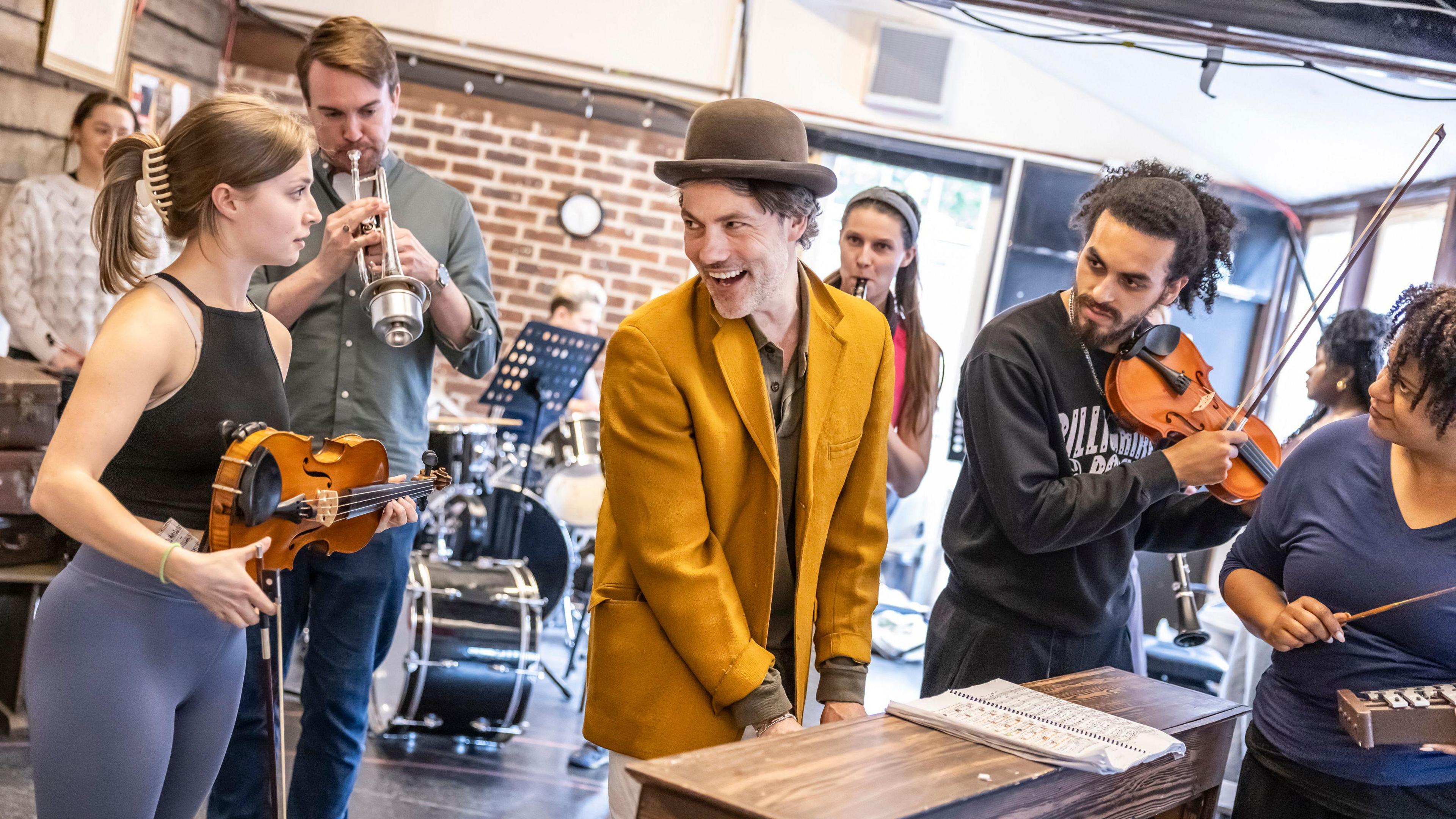 A lady playing the violin, a man playing a trumpet, a man playing the piano wearing a yellow blazer and bowler hat, and a man playing the violin