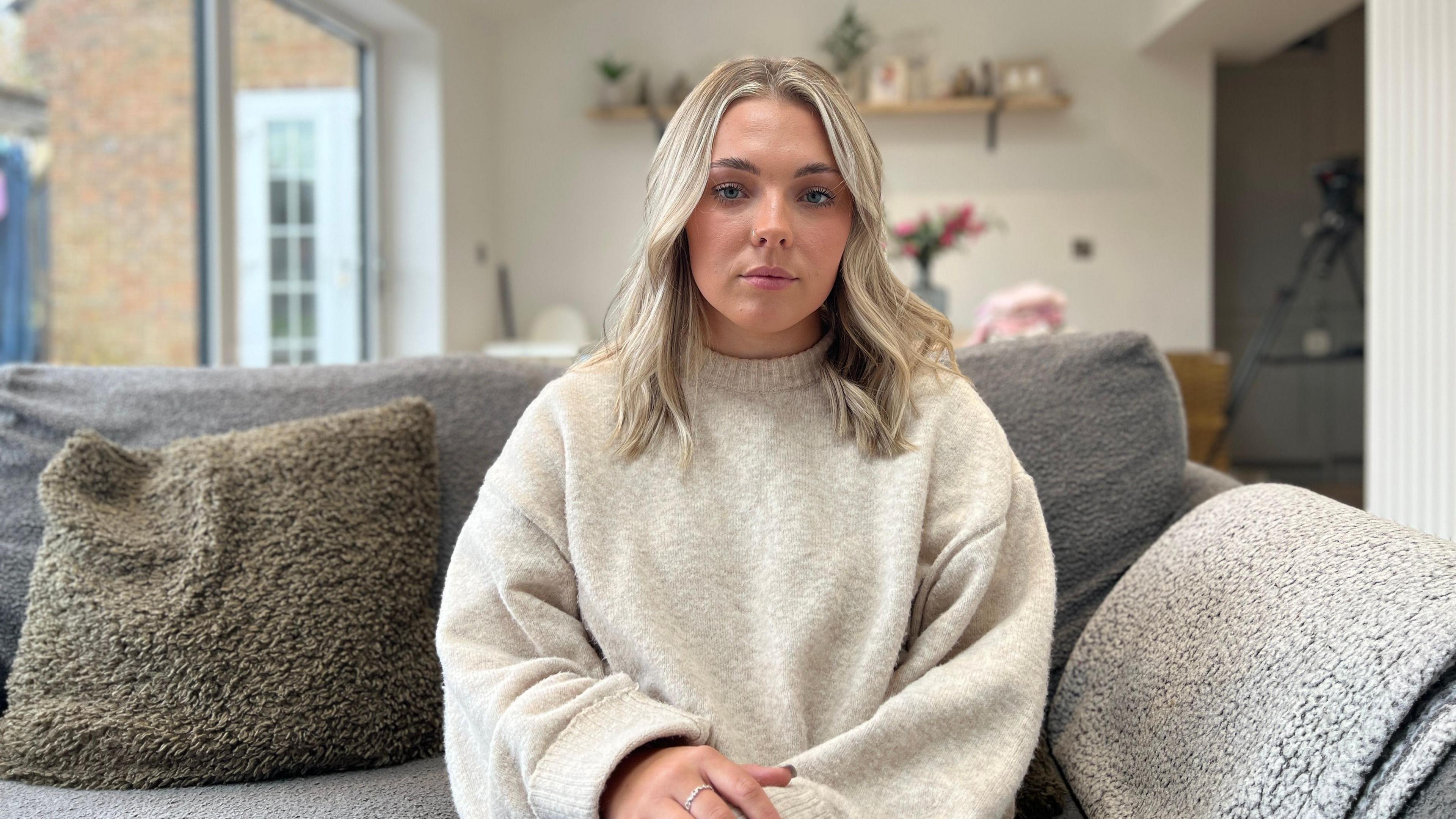 Megan is sitting on a grey sofa with a fleece blanket and fleece-covered cushion beside her. She has wavy blond hair and is wearing a cream coloured high neck knit jumper. Her expression is neutral and in the background you can see the top of a dining table with flowers in a vase on it and shelves with photos and knick knacks on above that