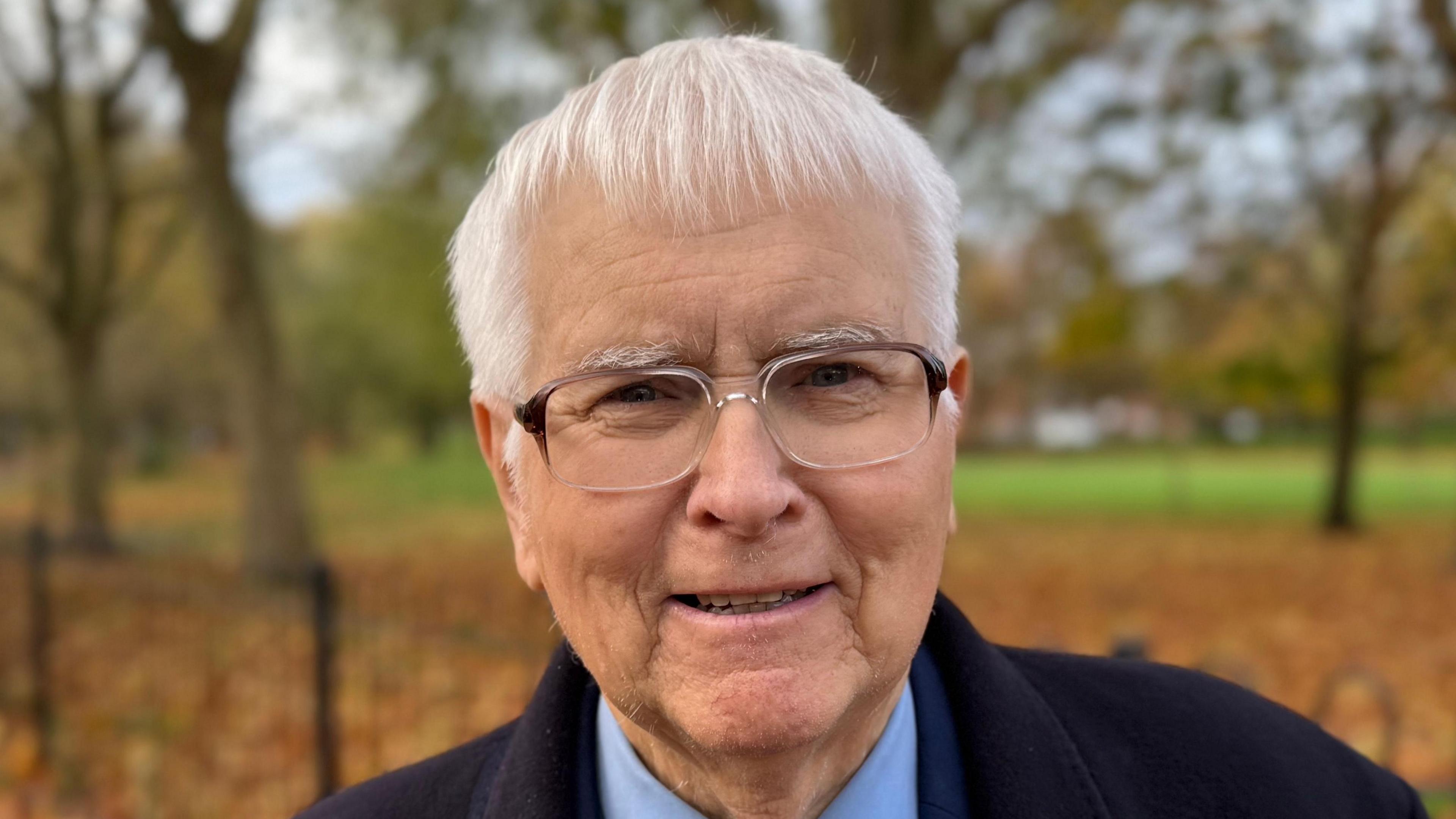 A man - Sir Bob Russell - smiling straight at the camera. He is wearing a black coat over a navy suit and is standing in a park.