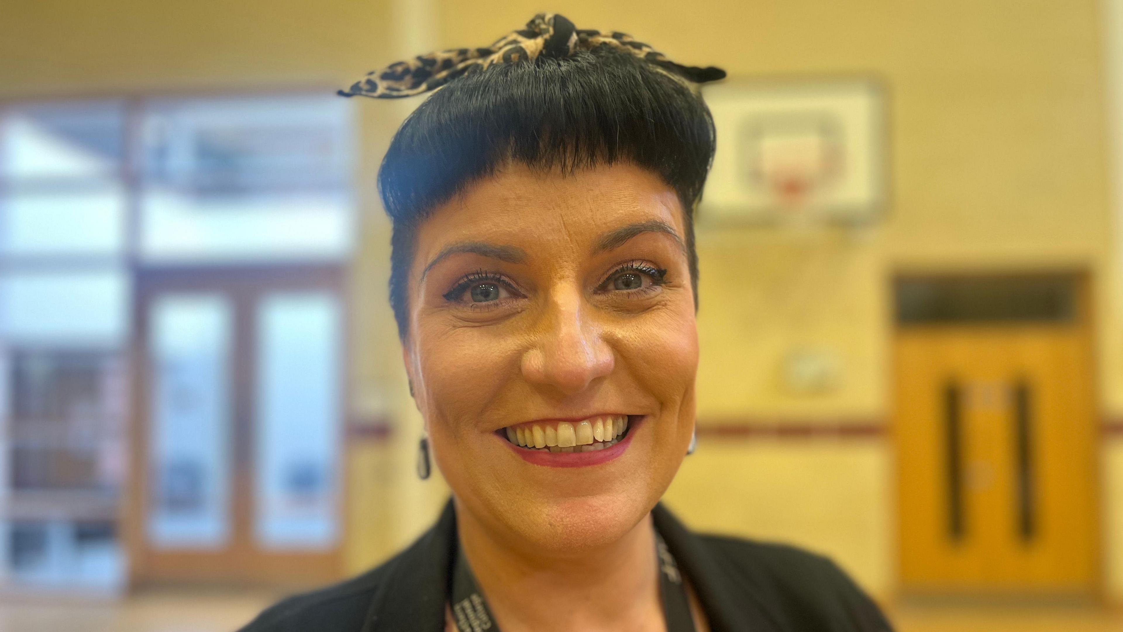 Teresa McBride is smiling into the camera. Shes standing in a gym in a school. There's a basketball hoop behind her. She is wearing a leopard print hairband.