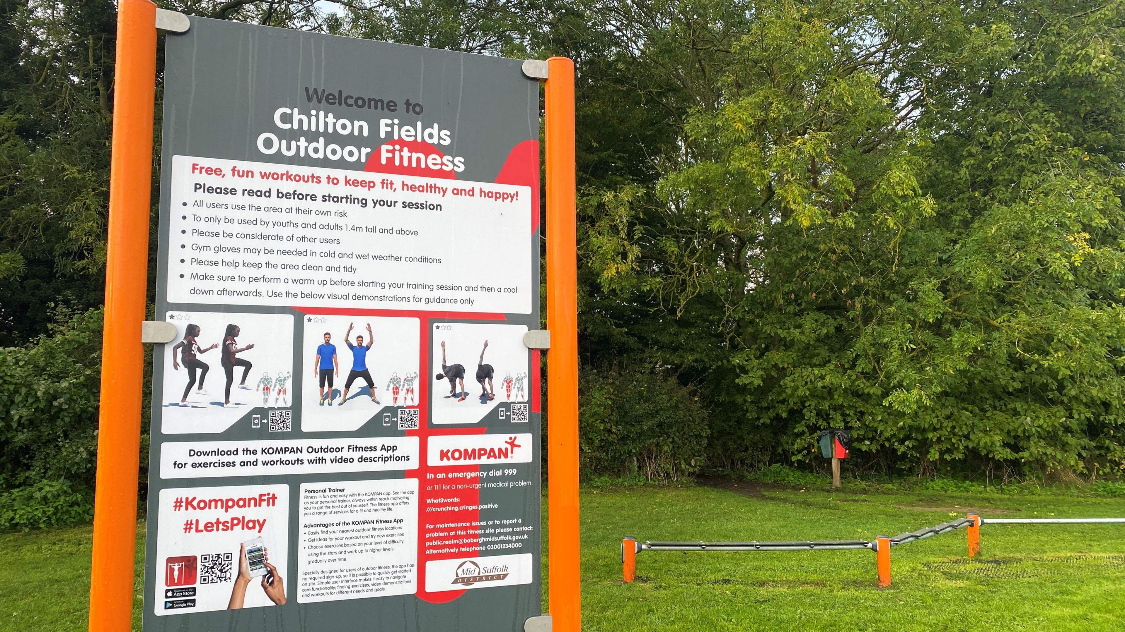 A grey sign attached to orange posts which reads Welcome to Chilton Fields Outdoor Fitness. It is in a field with trees in the background. 