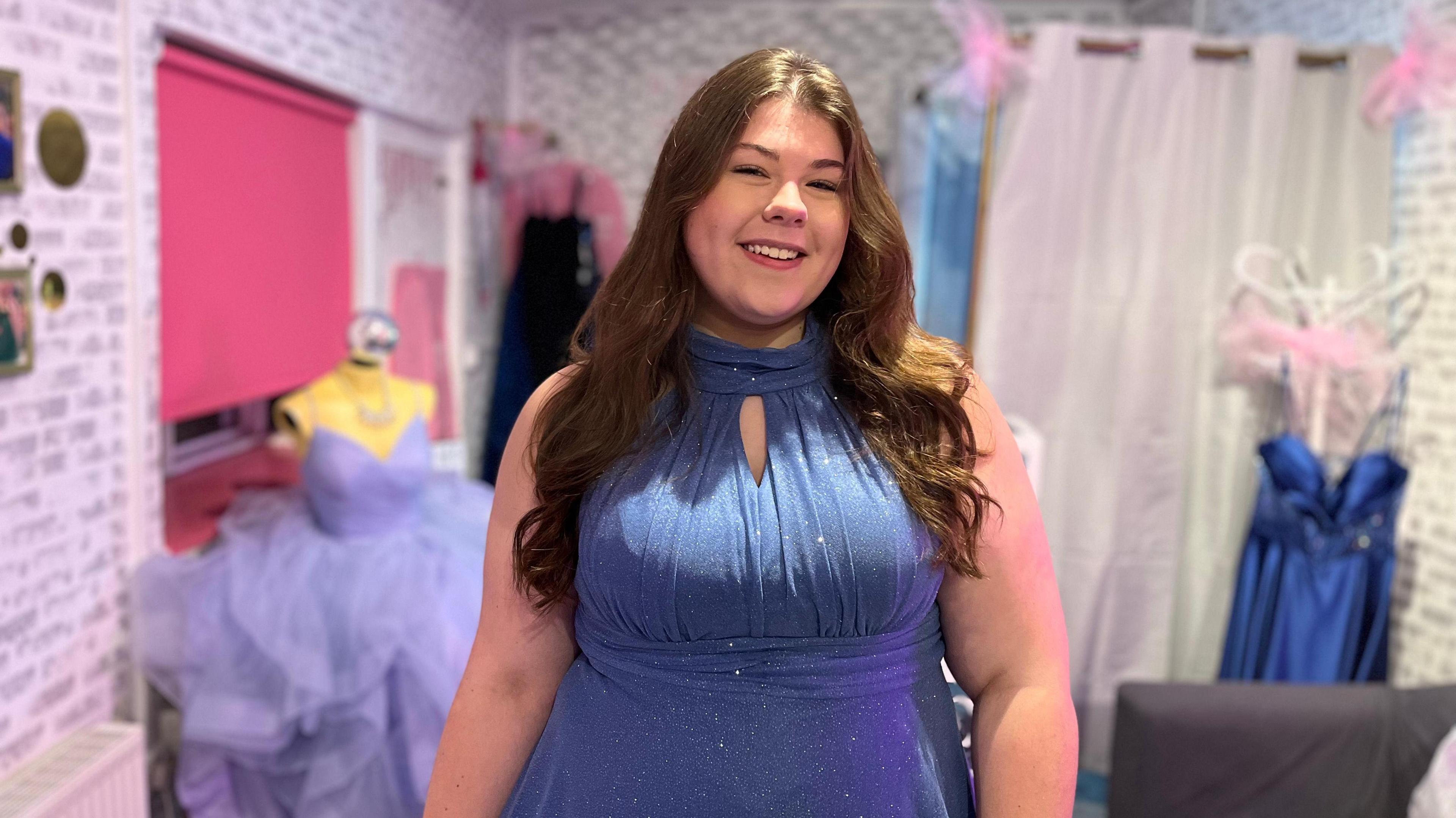 A teenage girl stands in a formal dress shop wearing a light blue sparkly halterneck evening dress. She has long brown wavy hair and is smiling. There is a strappy lilac ball gown on a mannequin in the background. 