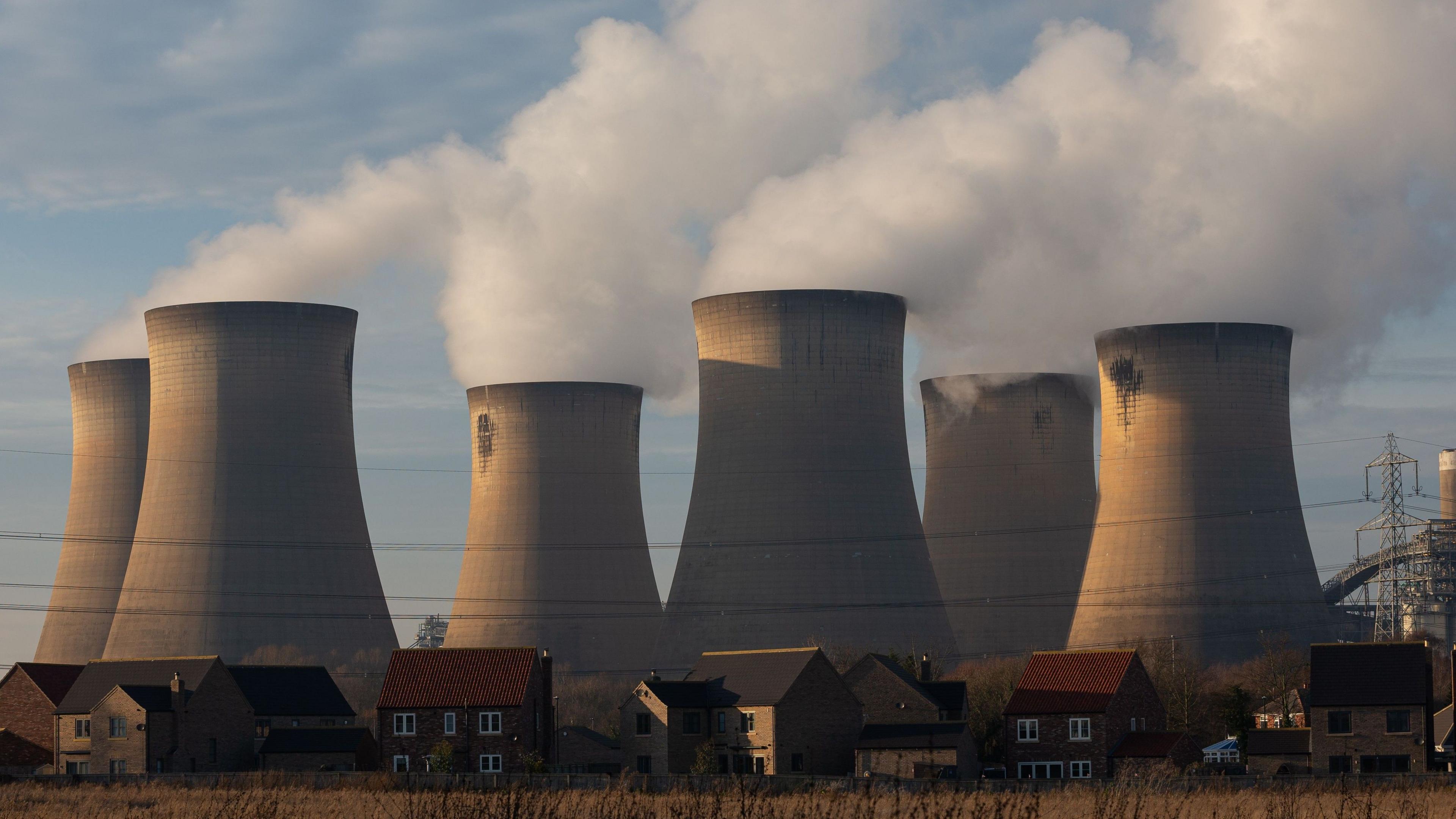 Draw power station. The photo shows smoke billowing out of six stout chimneys. There are a row of houses and a number of electricity cables in the foreground.